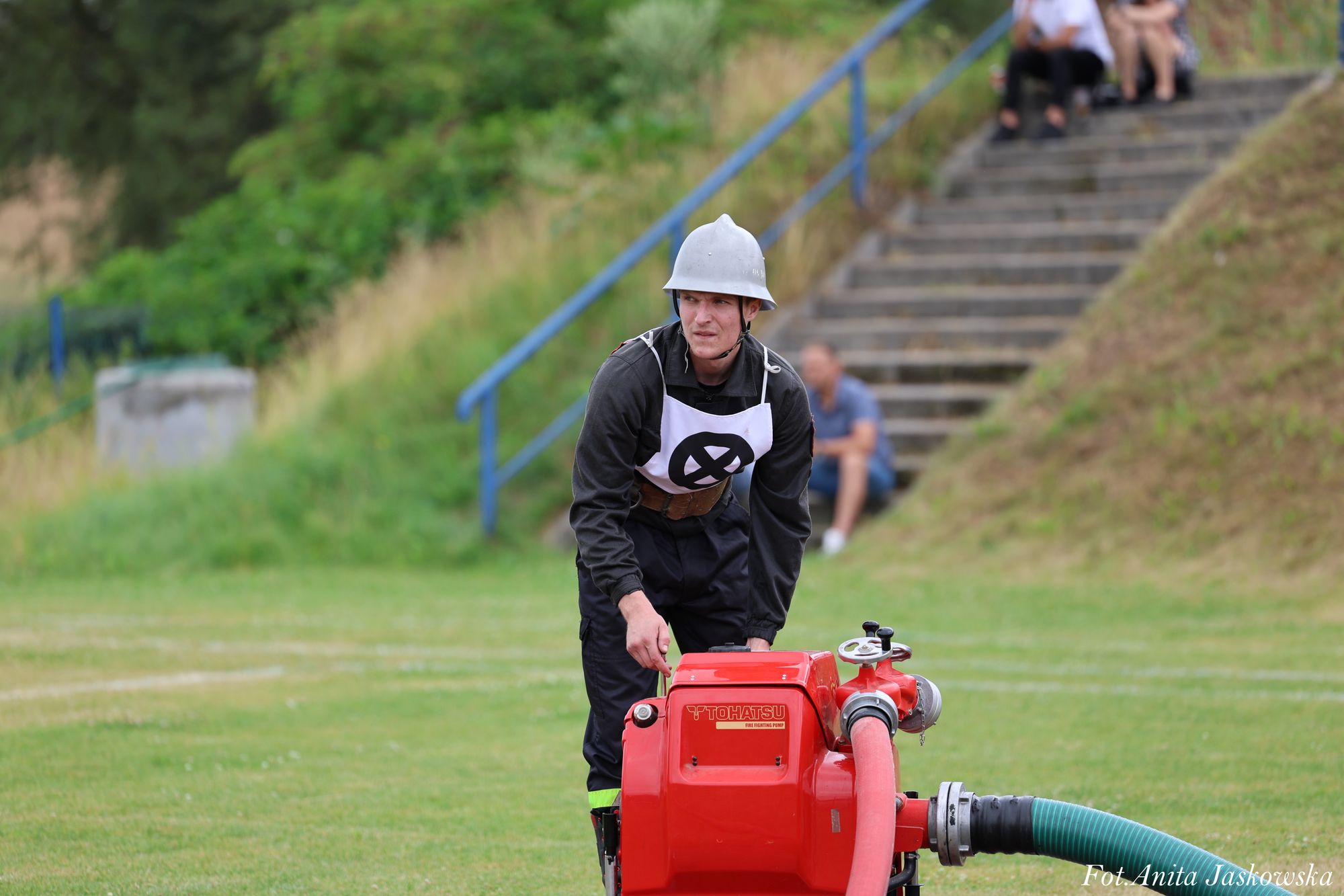 Mężczyzna w czarnym ubraniu ochronnym i szarym hełmie pochylony nad czerwonym urządzeniem z podłączonymi wężami na zielonej trawie, w tle schody z niebieską poręczą.