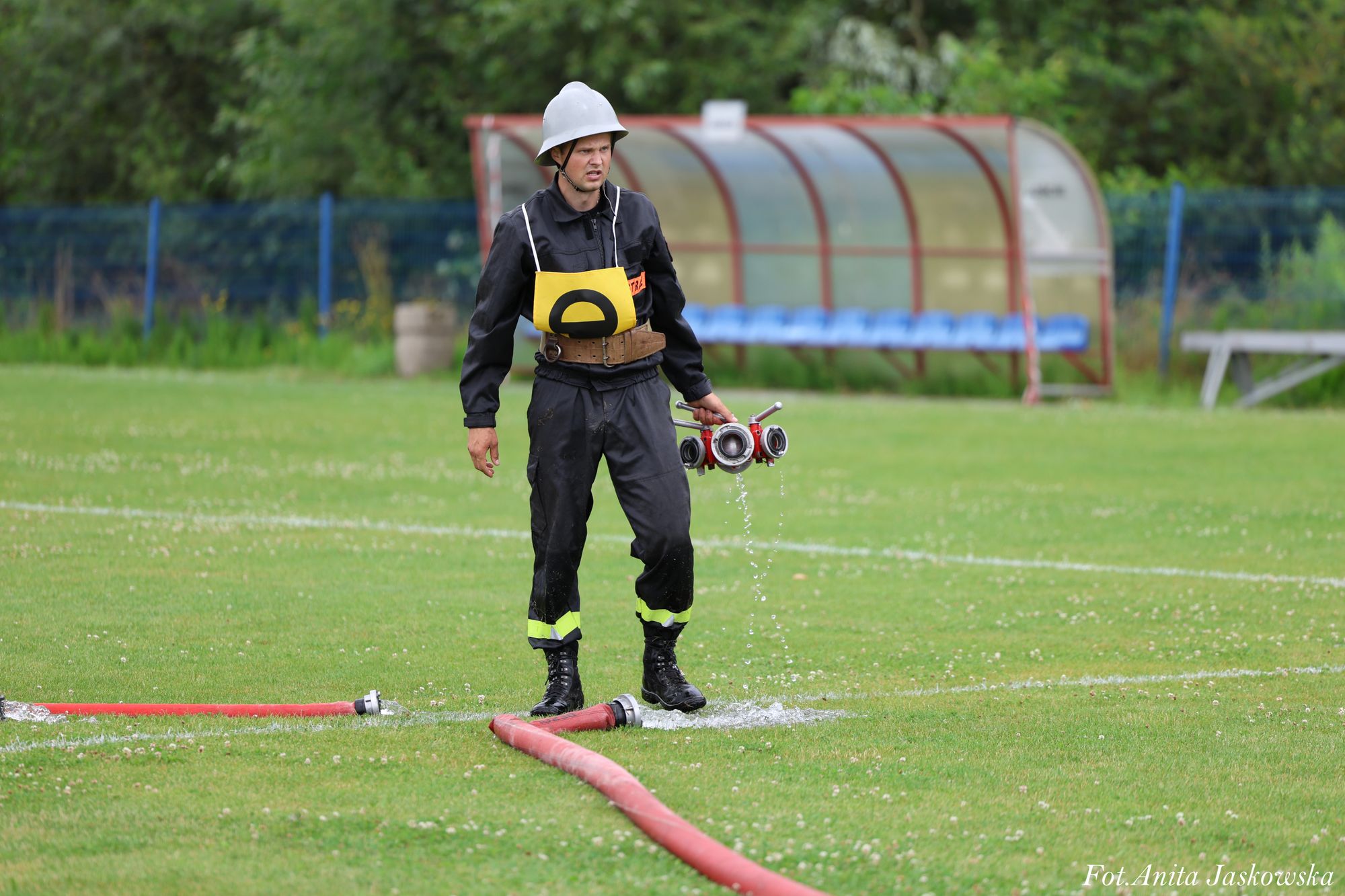 Mężczyzna w czarnym ubraniu strażaka i szarym hełmie stoi na zielonej trawie, trzyma czerwony rozdzielacz strażacki z cieknącą wodą, w tle zadaszone ławki i ogrodzenie.