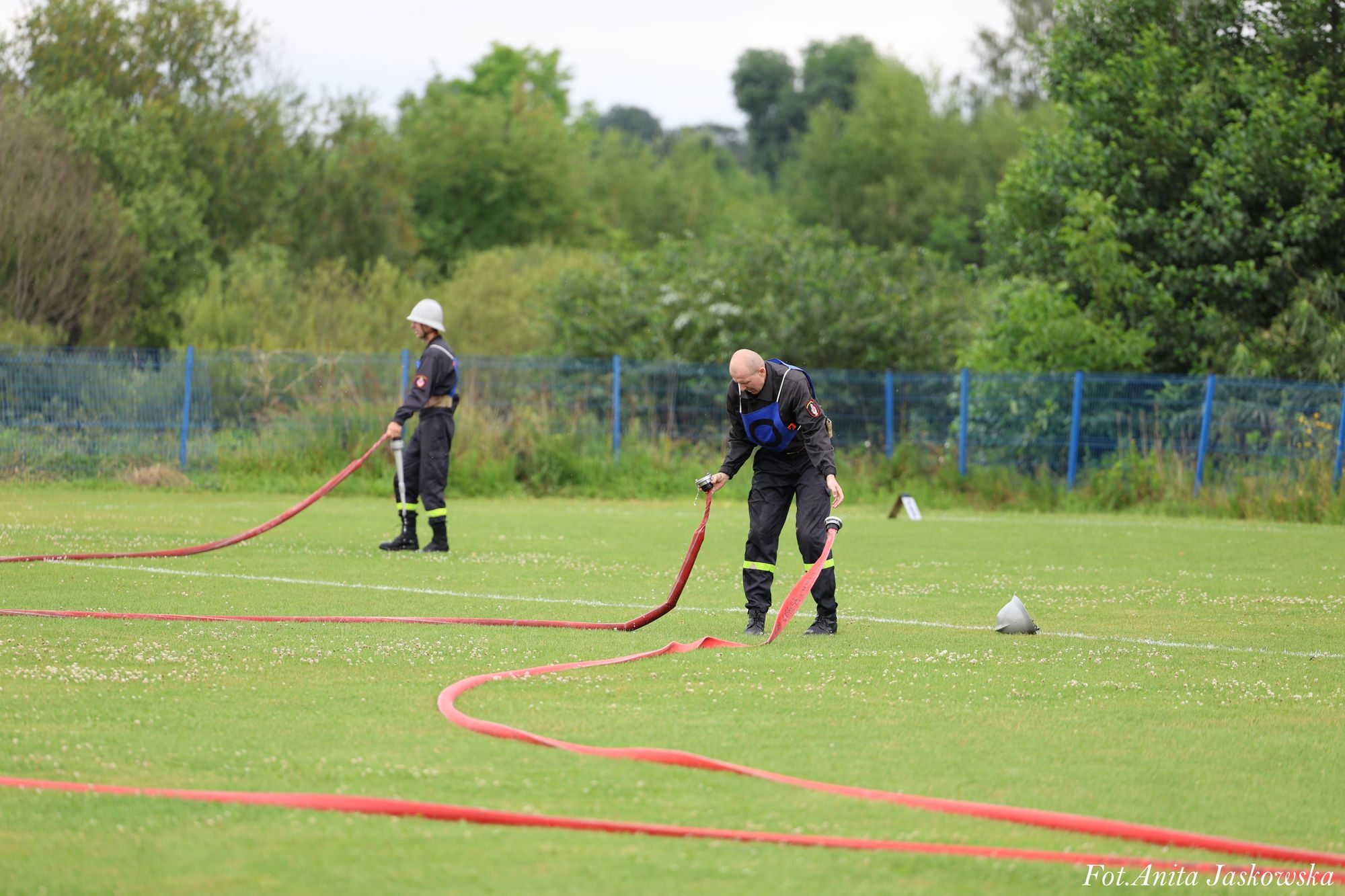 Dwóch mężczyzn w czarnych ubraniach z żółtymi odblaskami rozwija długie czerwone węże na zielonym trawiastym boisku, w tle zieleń i niebieski płot.
