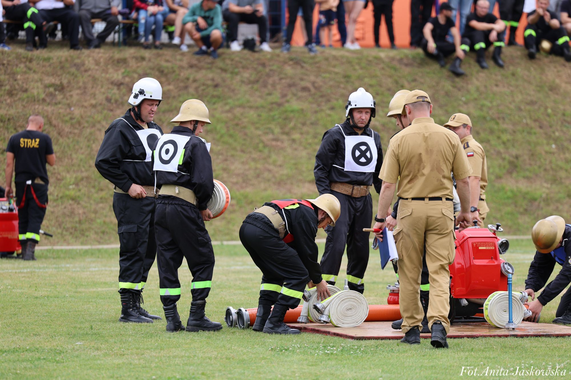Grupa strażaków w czarnych mundurach i kaskach, zwijające białe węże strażackie na zielonej trawie, obok nich dwie osoby w beżowych mundurach.