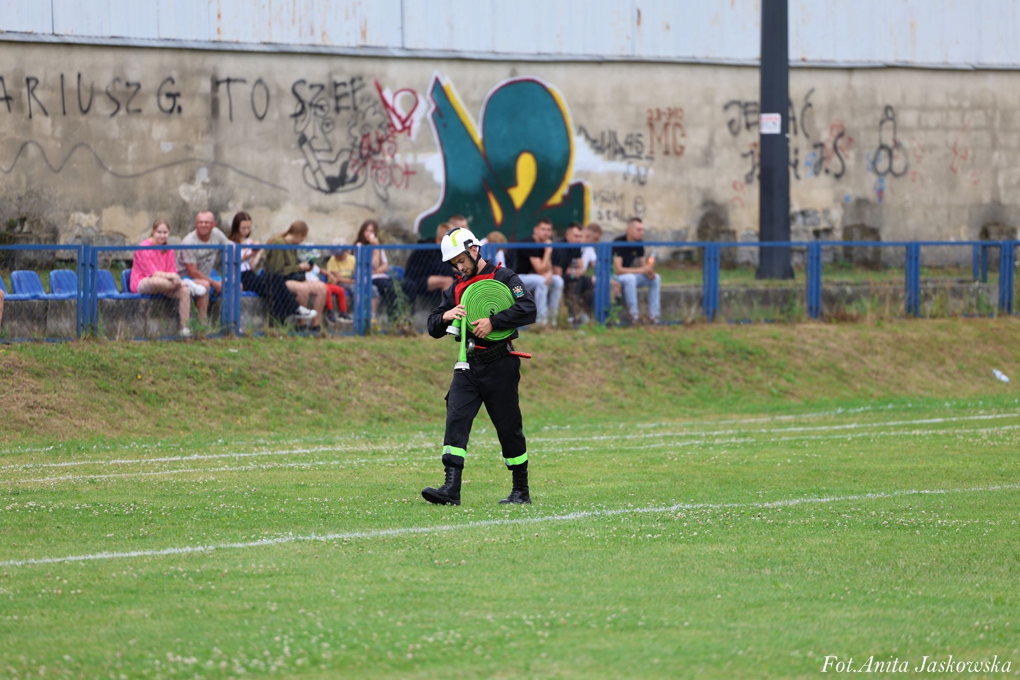 Strażak w czarnym ubraniu z odblaskami i białym hełmem niesie zielony zwinięty wąż na tle zielonej trawy i murku z graffiti oraz kilku siedzących osób.