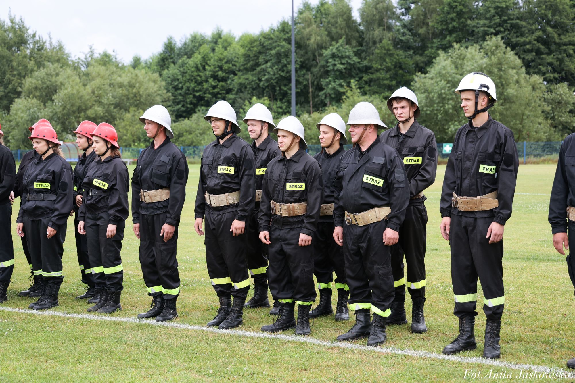 Grupa strażaków w czarnych mundurach z żółtymi naszywkami "STRAŻ", czerwonymi i białymi hełmami, stojąca na zielonym trawiastym boisku, w tle drzewa.