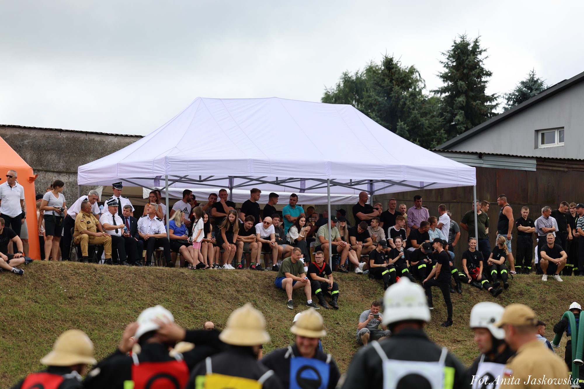 Grupa ludzi siedzących i stojących pod białym namiotem na tle trawy i budynków, na pierwszym planie osoby w hełmach strażackich.
