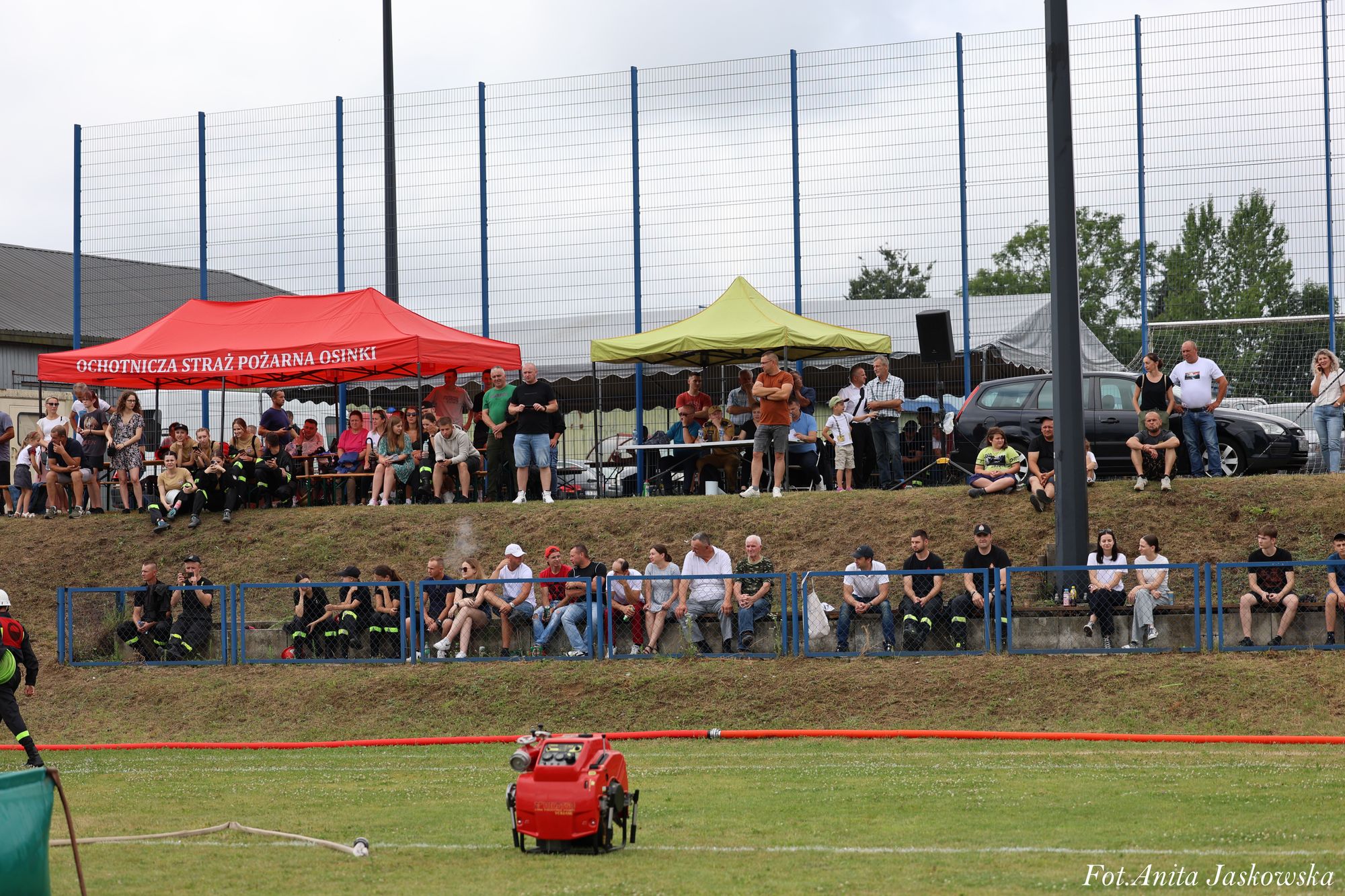 Ludzie siedzący i stojący na trawiastym zboczu z czerwonym i zielonym namiotem, niebieskim ogrodzeniem, zieloną trawą i czerwonym sprzętem strażackim na pierwszym planie.