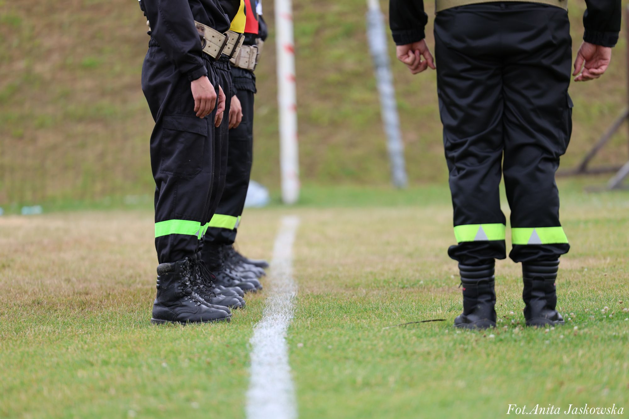 Cztery osoby w czarnych spodniach z odblaskowymi paskami i czarnych butach stojące na trawiastej powierzchni przy białej linii.
