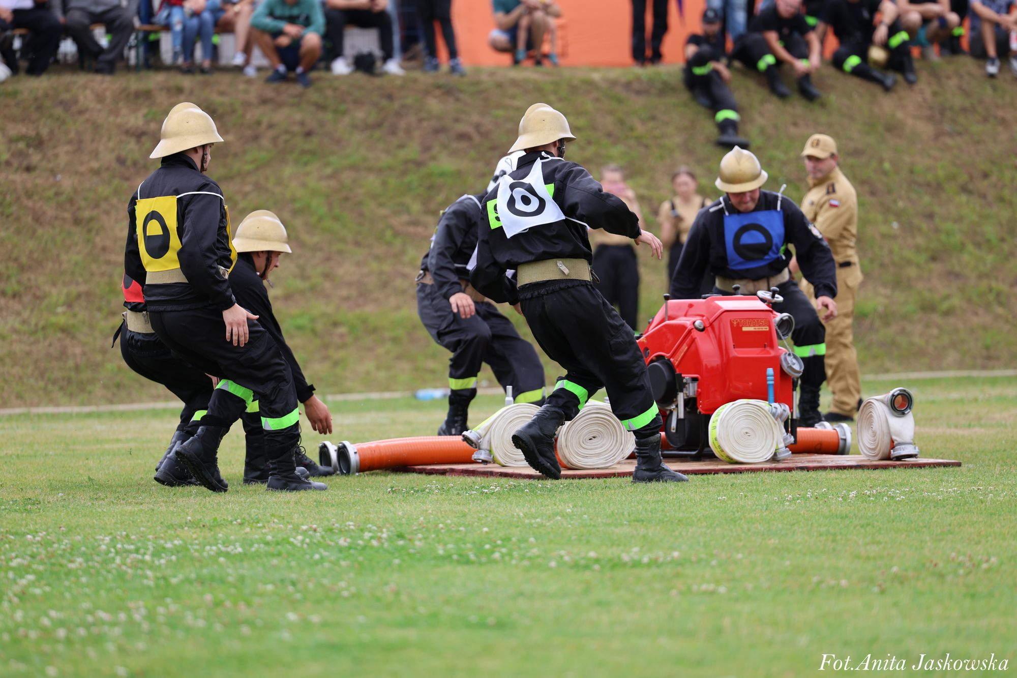 Grupa strażaków w czarnych mundurach z żółtymi i niebieskimi oznaczeniami, beżowe hełmy, rozkładająca sprzęt strażacki na zielonej trawie, w tle widzowie na skarpie.