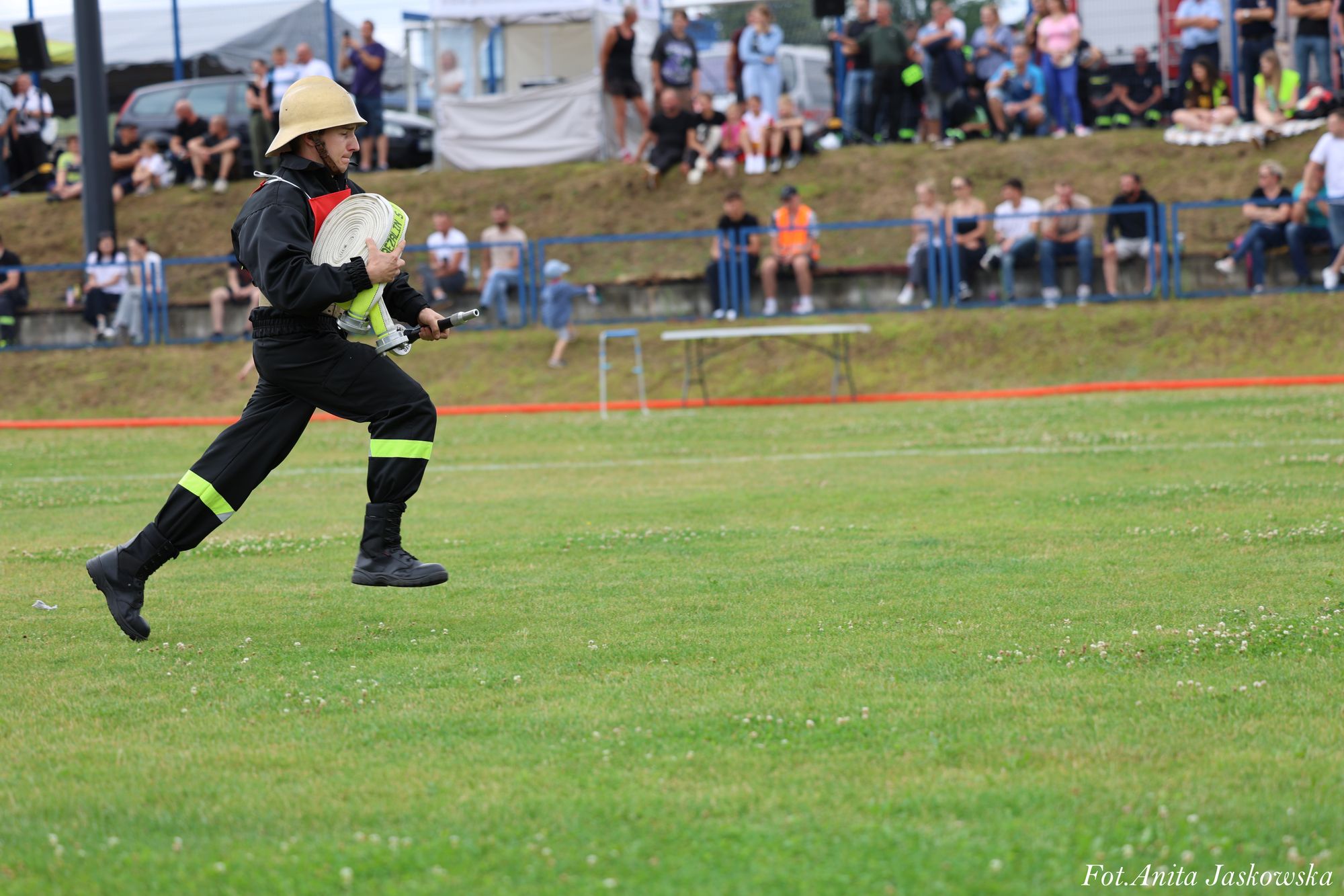Strażak w czarnym ubraniu z odblaskowymi paskami i żółtym hełmem biegnie z białym zwiniętym wężem strażackim na zielonej trawie, w tle ludzie na widowni.