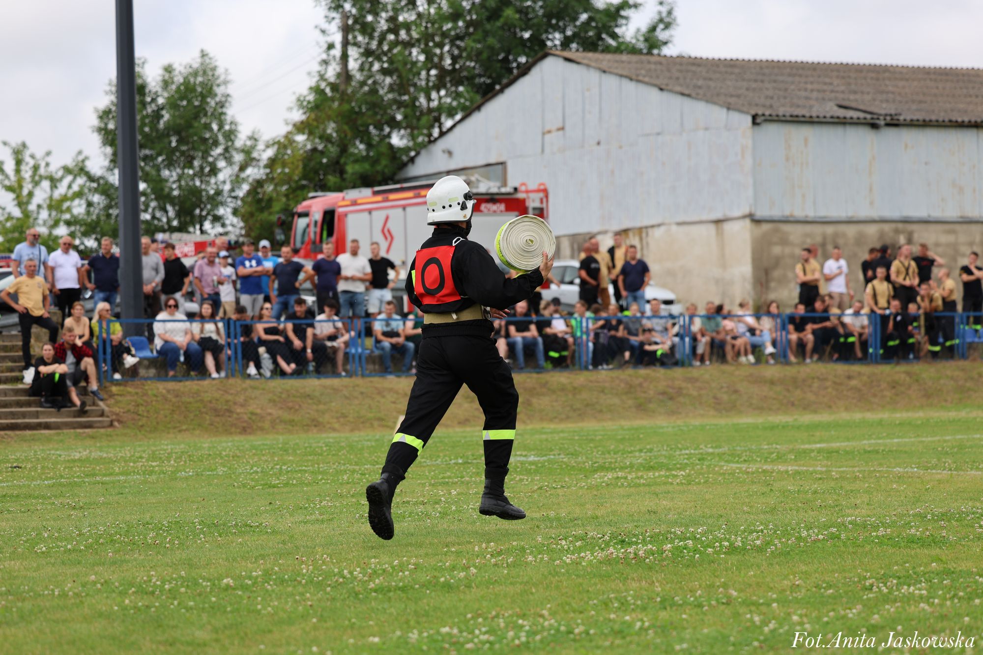 Strażak w czarnym ubraniu z żółtymi odblaskami i białym hełmem biegnie po zielonym boisku, trzymając zwinięty wąż strażacki, w tle widoczna publiczność i wóz strażacki.