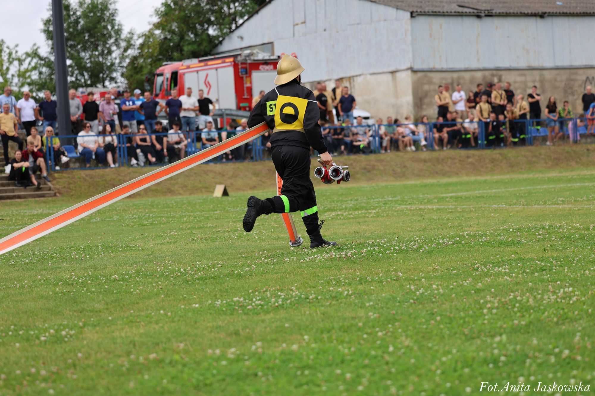 Strażak w czarnym ubraniu z żółtym numerem 0 na plecach biegnie po zielonym trawniku, trzymając czerwony wąż pożarniczy i sprzęt, w tle tłum ludzi i wóz strażacki.