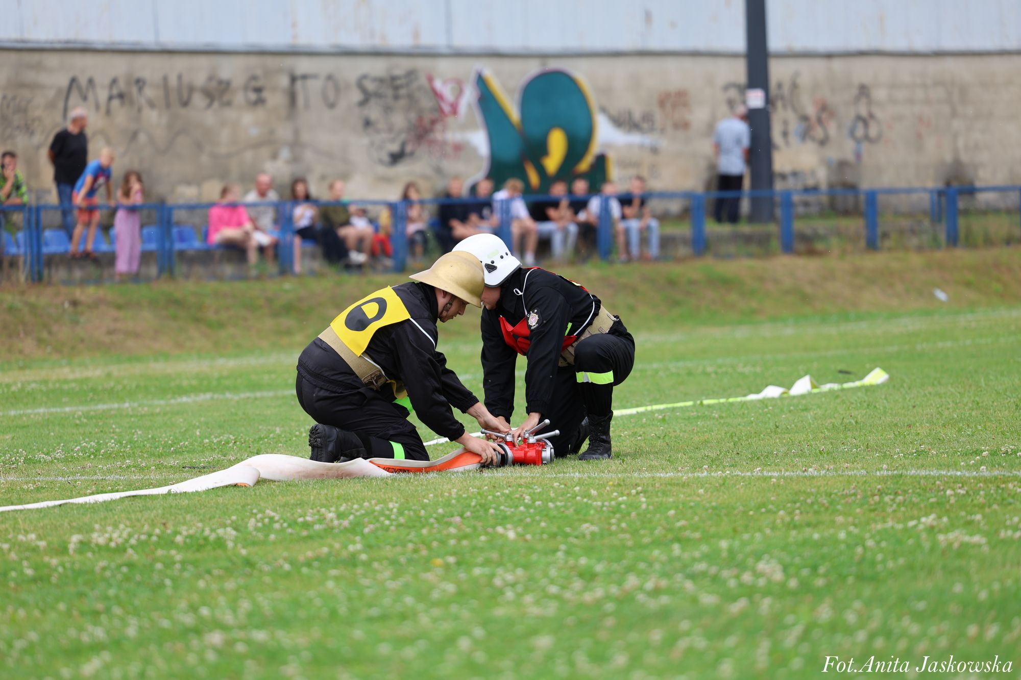 Dwóch strażaków w hełmach i czarnych mundurach z żółtymi i czerwonymi oznaczeniami klęczy na zielonej trawie, łącząc białe i pomarańczowe węże strażackie, w tle rozmyta grupa ludzi i szary mur z graffiti.