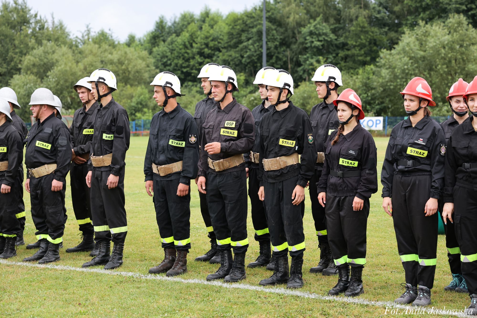 Grupa strażaków w czarnych mundurach z neonowymi wstawkami i kaskami białymi oraz czerwonymi, stojących na zielonej trawie, za nimi drzewa.