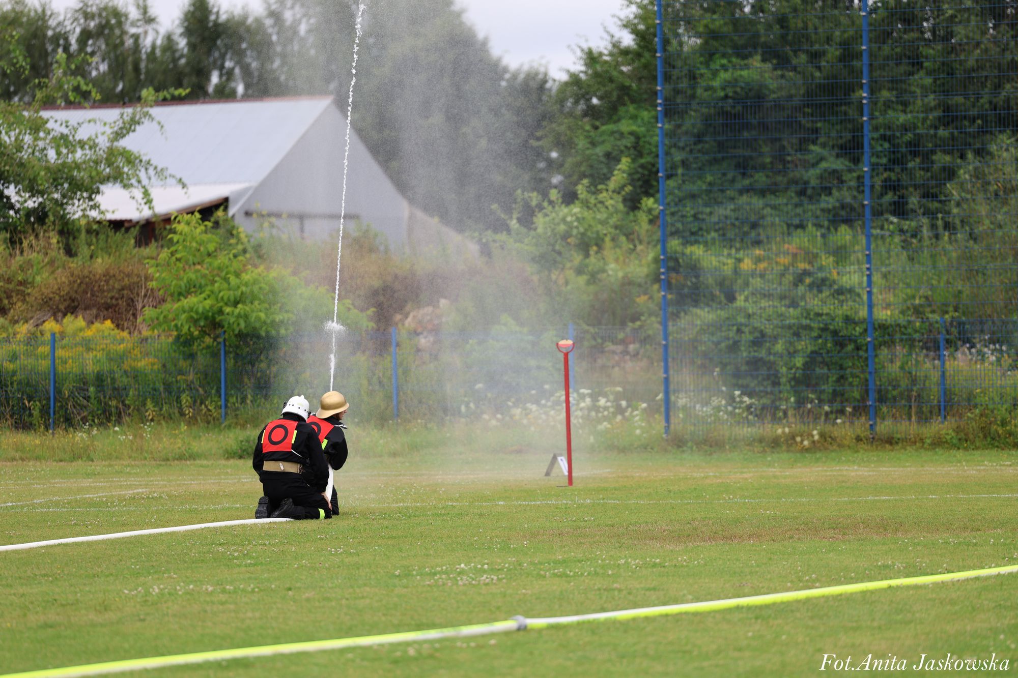 Dwóch strażaków w czarnych ubraniach z czerwonymi oznaczeniami, jeden w białym hełmie, drugi w złotym, klęczy na trawie obok strumienia wody z węża strażackiego.