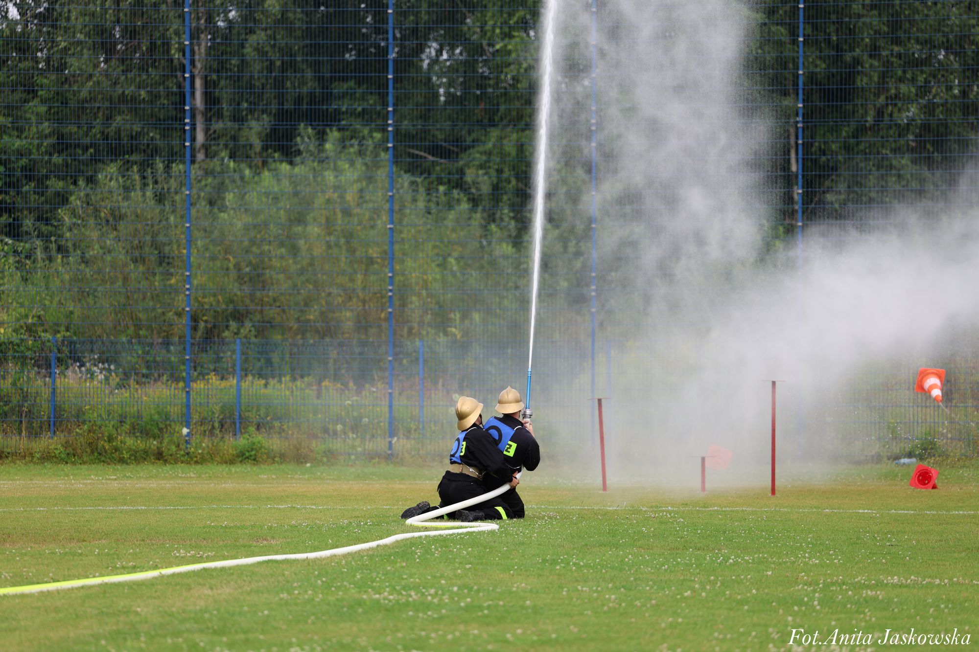 Dwóch strażaków w hełmach i ubraniach ochronnych klęczy na trawie, trzymając wąż strażacki skierowany pionowo, z którego leci strumień wody. Tło to zielone drzewa i niebieska siatka.