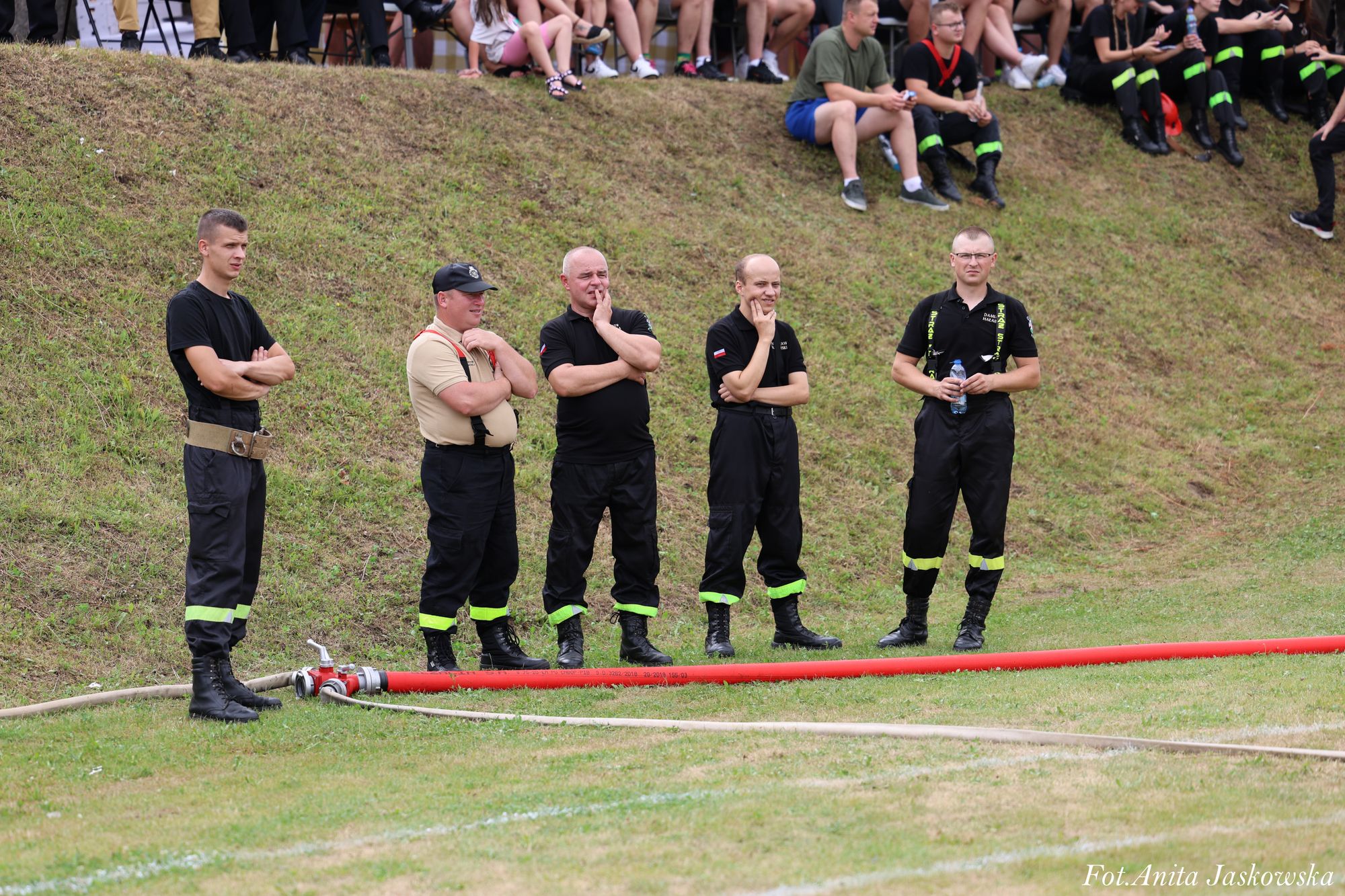 Pięciu mężczyzn w czarnych i beżowych ubraniach strażackich stoi na trawie obok czerwonego węża strażackiego, za nimi widać nachylone wzgórze z siedzącymi ludźmi.