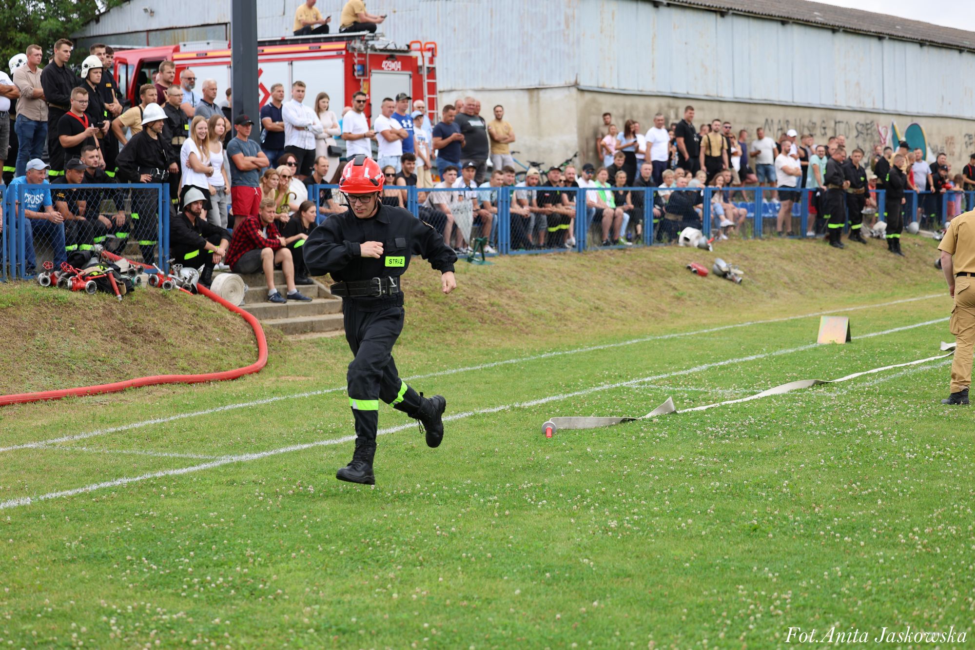 Mężczyzna w czarnym ubraniu strażackim i czerwonym hełmie biegnie po zielonym boisku, za nim tłum ludzi i czerwony wóz strażacki.
