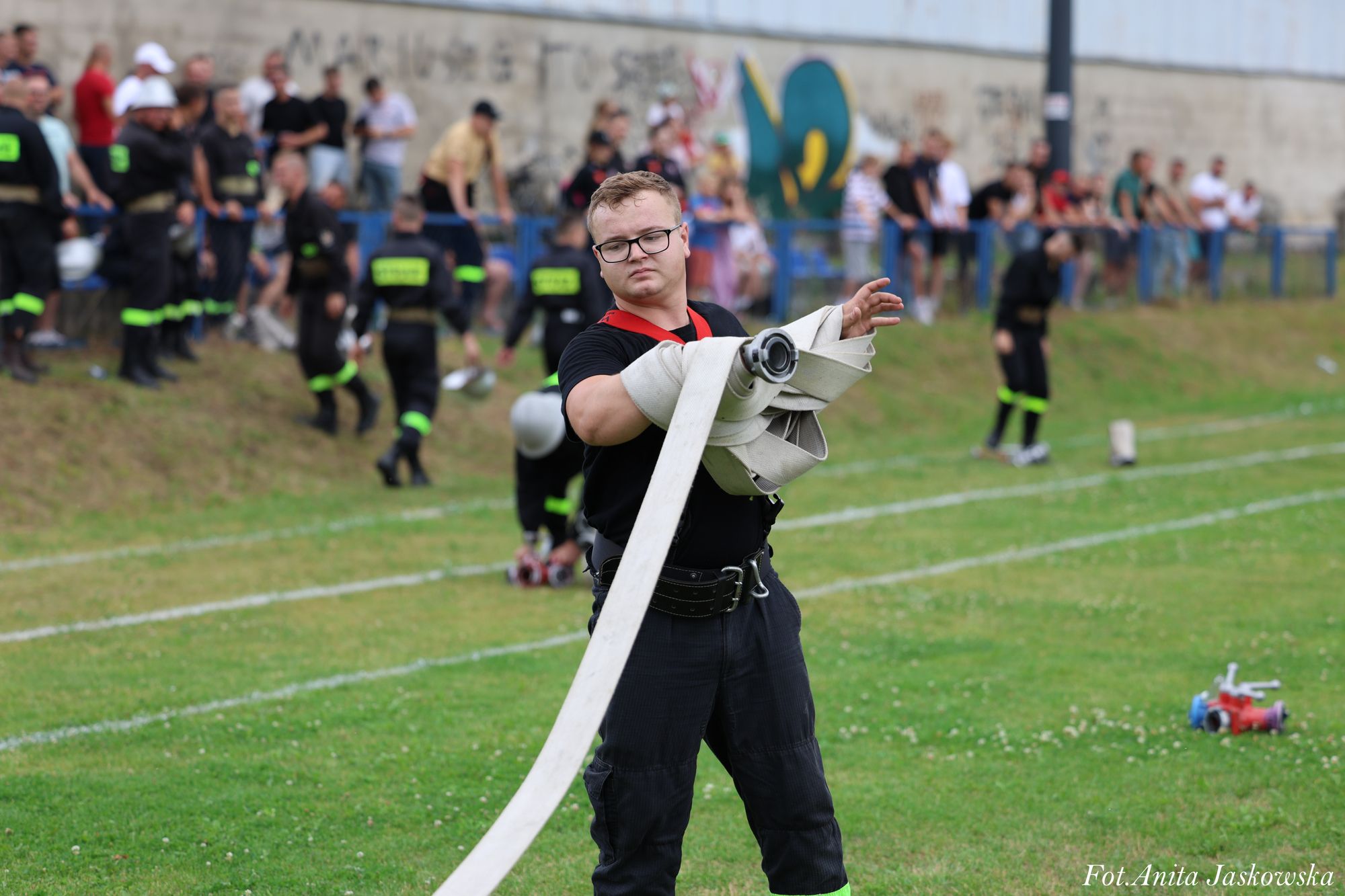 Mężczyzna w czarnym ubraniu i okularach rozwija biały wąż strażacki na zielonej trawie, w tle grupa ludzi i mur z graffiti.
