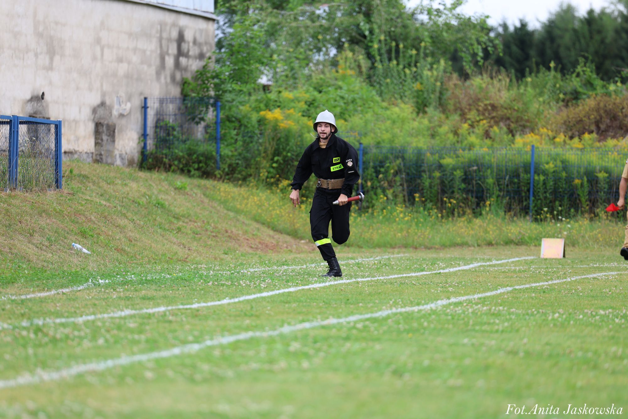 Mężczyzna w czarnym stroju strażackim z białym hełmem biegnie po trawiastym boisku z białymi liniami, trzymając krótki wąż strażacki. Tło: zarośla, budynek, ogrodzenie.