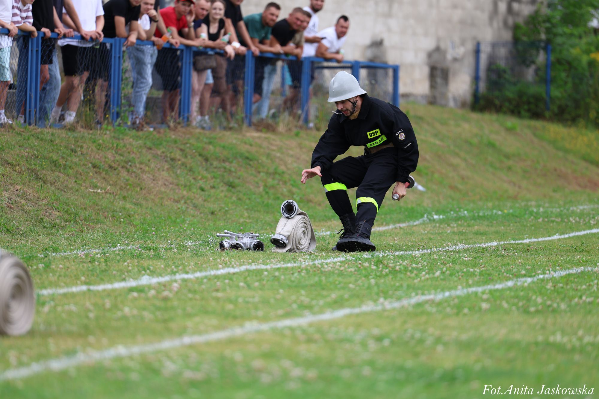 Mężczyzna w czarnym uniformie strażaka z odblaskowymi paskami i hełmem kuca na trawie obok zwiniętego białego węża strażackiego, w tle tłum ludzi za niebieskim ogrodzeniem.