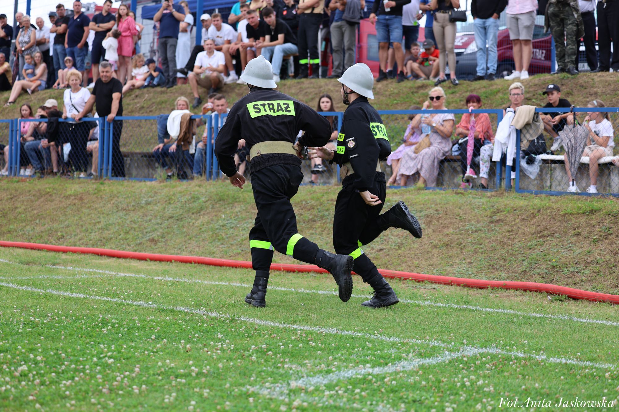 Dwóch strażaków w czarnych mundurach i hełmach biegnie po zielonej trawie, w tle grupa siedzących i stojących osób za niebieskim ogrodzeniem.