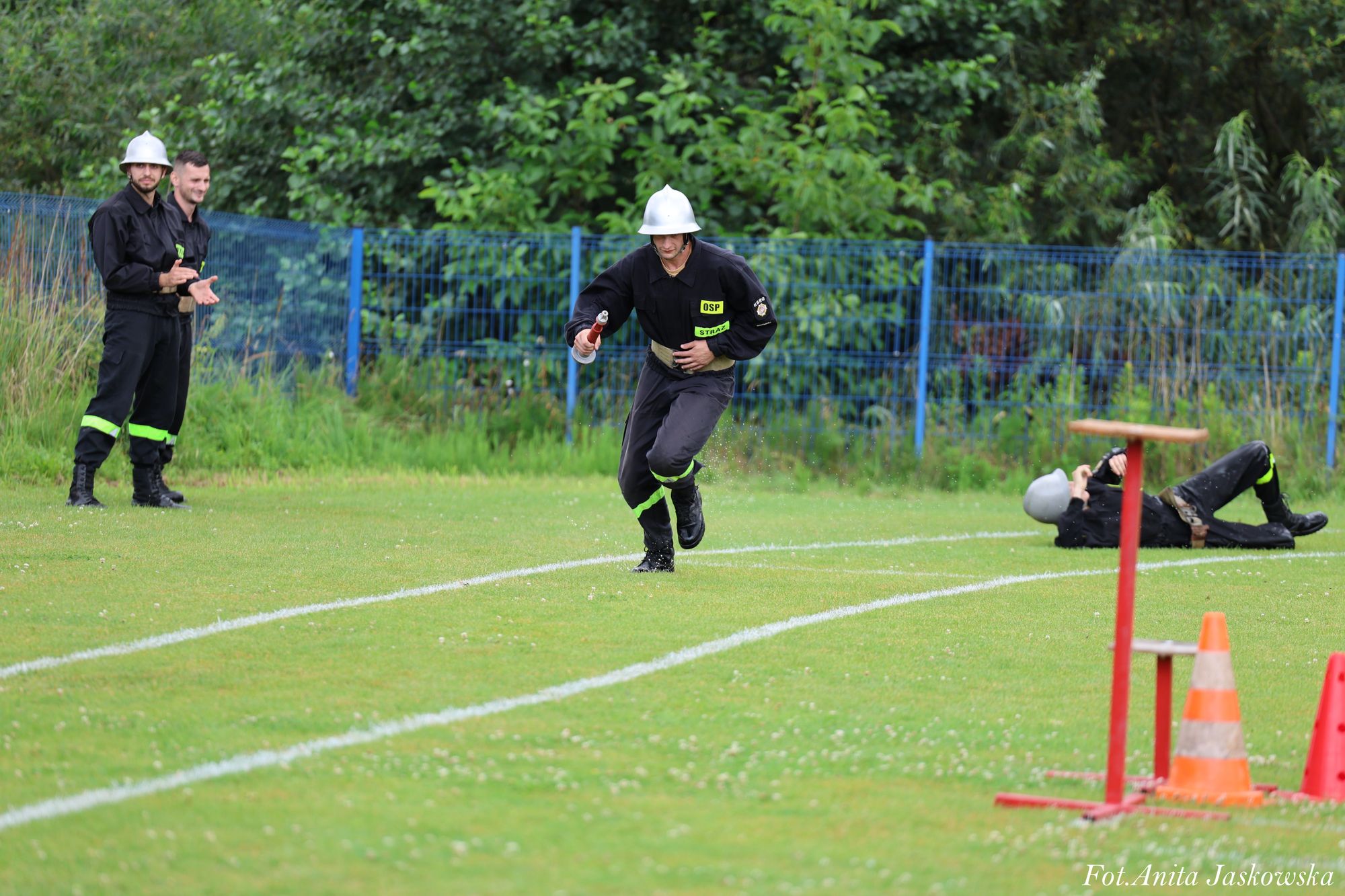 Czterech mężczyzn w czarnych mundurach strażackich na zielonym boisku, jeden biegnie z wężem, jeden leży na trawie, dwóch stoi obok siebie przy płocie.