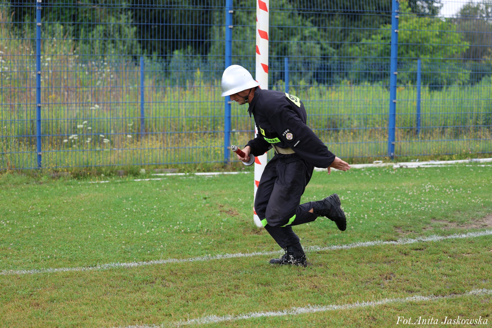 Strażak w czarnym mundurze i białym hełmie biegnący po trawiastym boisku obok niebieskiego ogrodzenia i słupka z biało-czerwonymi paskami.