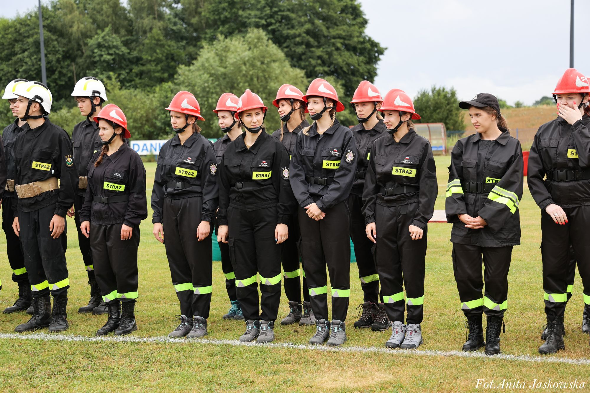 Grupa młodych strażaków i strażaczek w czarnych mundurach z żółtymi odblaskami, czerwonych i białych hełmach, stojąca na trawiastym boisku na tle zielonych drzew.