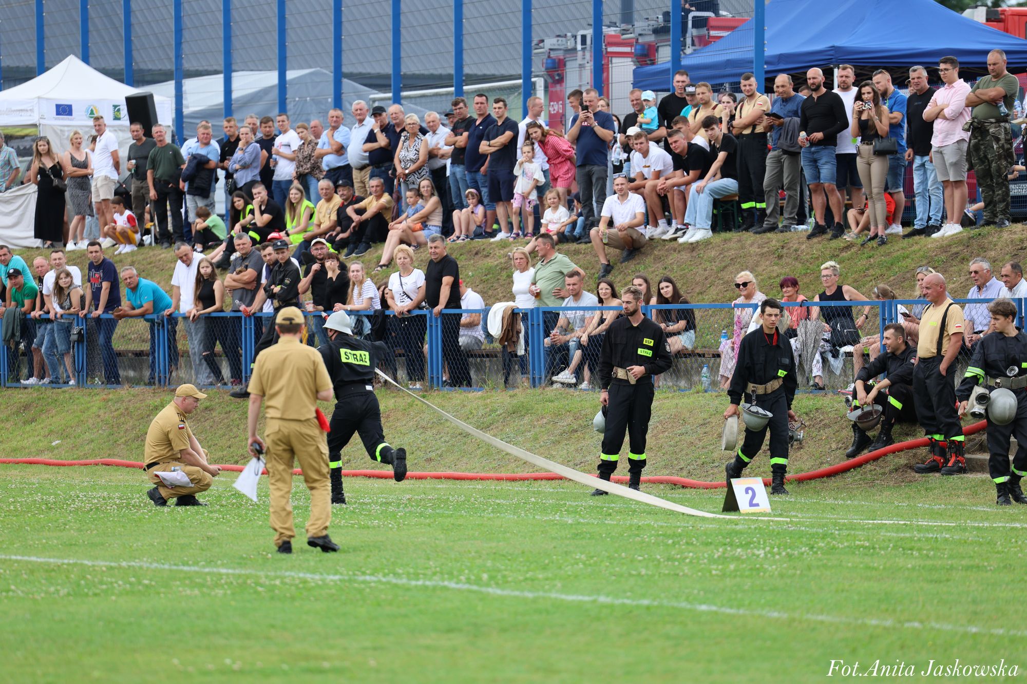 Grupa strażaków w czarnych mundurach z żółtymi napisami na zielonym tle na trawie, w tle tłum ludzi za niebieskim ogrodzeniem i namiot niebieski oraz biały.