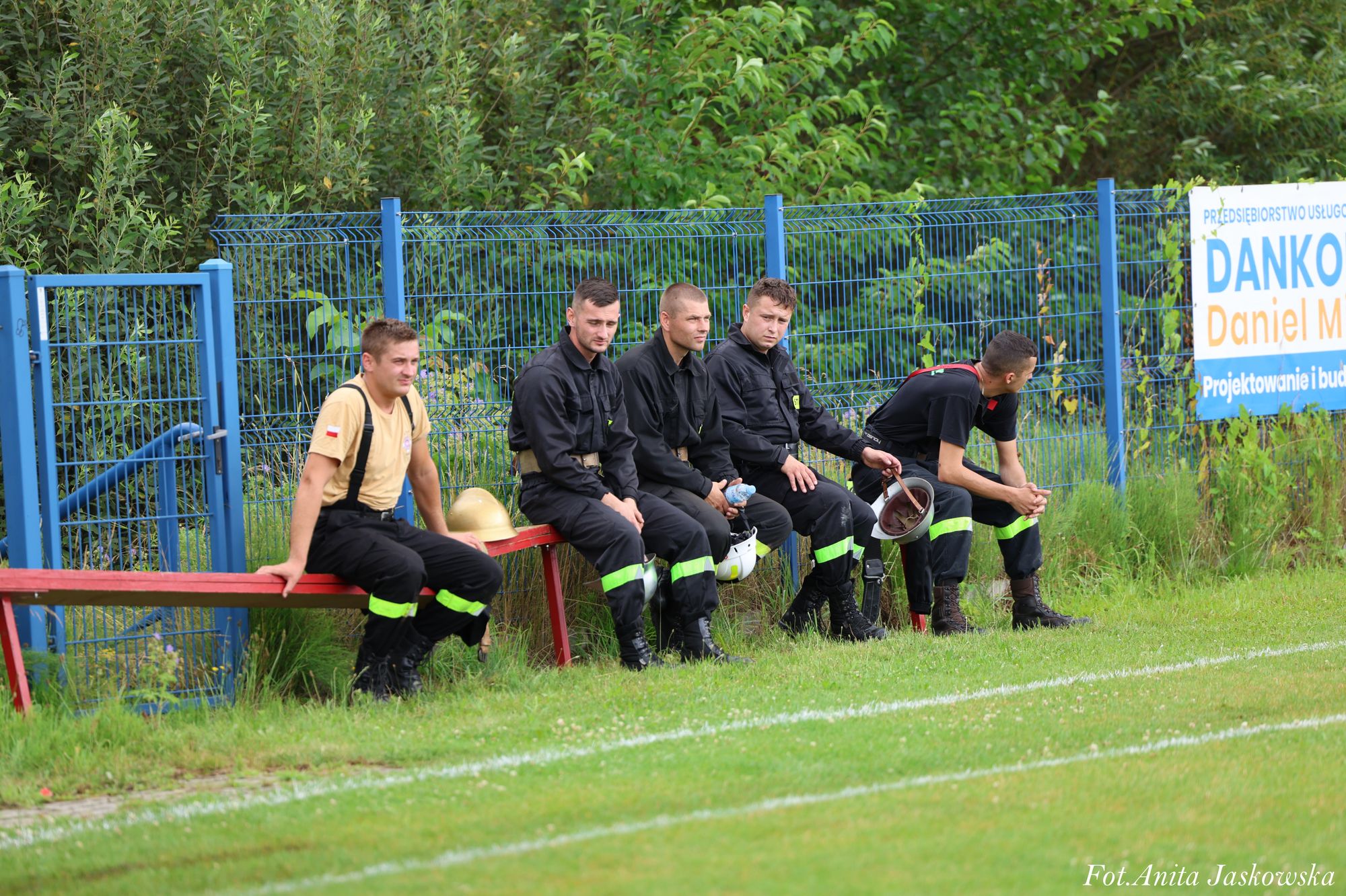 Pięciu mężczyzn w czarnych mundurach z odblaskowymi zielonymi pasami siedzi na czerwonej ławce na tle niebieskiego ogrodzenia i zielonych krzewów.