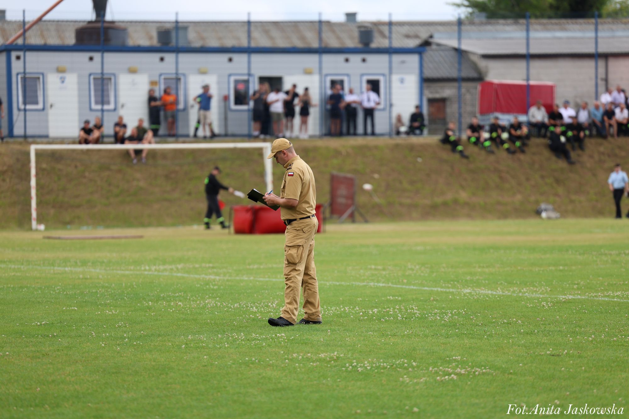 Mężczyzna w beżowym mundurze i czapce na zielonym boisku, w tle ludzie siedzący na skarpie i budynek z szarym dachem, biały bramkarz piłkarski.