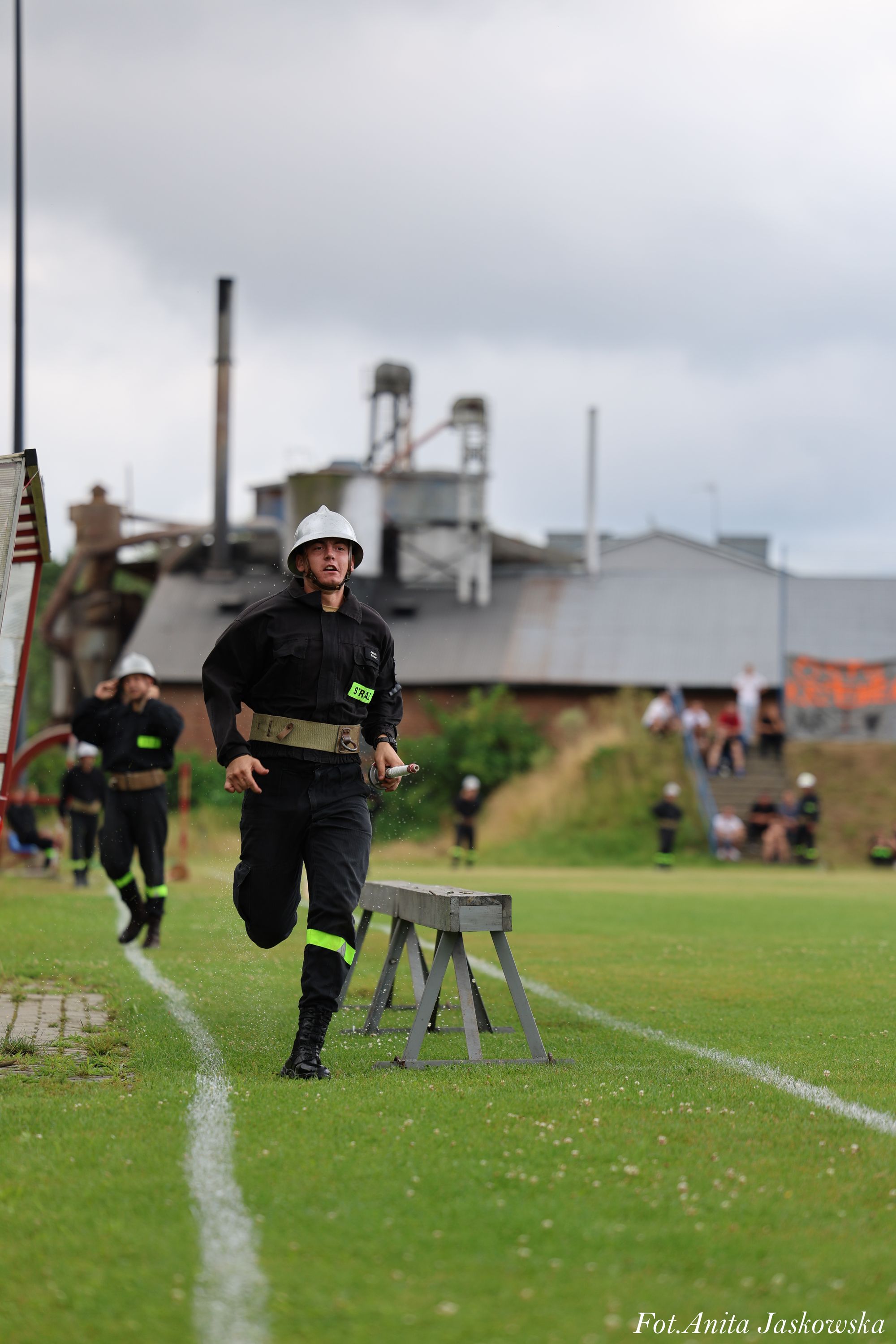 Mężczyzna w czarnym mundurze strażackim i srebrnym hełmie biegnie po zielonej trawie obok drewnianej ławki, w tle inne osoby w podobnych mundurach i przemysłowe budynki.