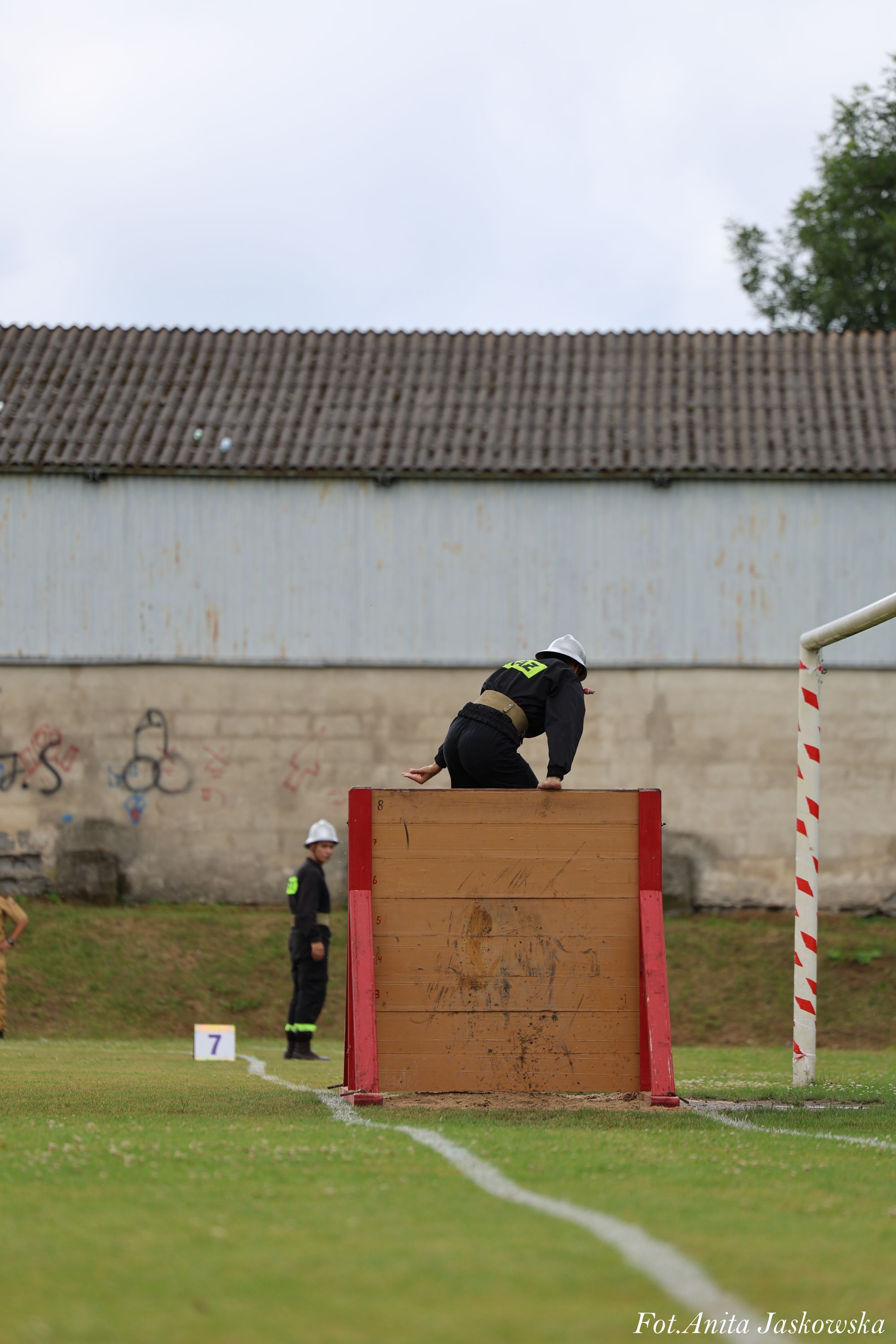 Osoba w czarnym stroju i białym hełmie pokonuje drewnianą przeszkodę z czerwonymi bokami na zielonym boisku sportowym. W tle mur z graffiti i inna osoba w podobnym stroju.