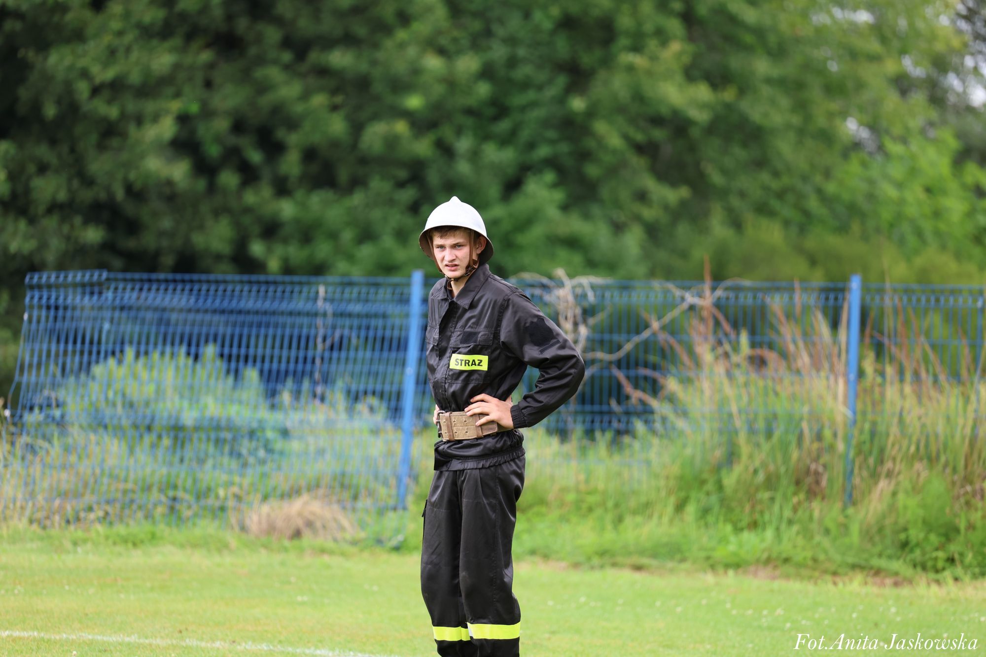 Młody mężczyzna w czarnym uniformie strażackim z żółtymi odblaskami i białym hełmem, stojący na zielonej trawie przed niebieskim ogrodzeniem i zaroślami.