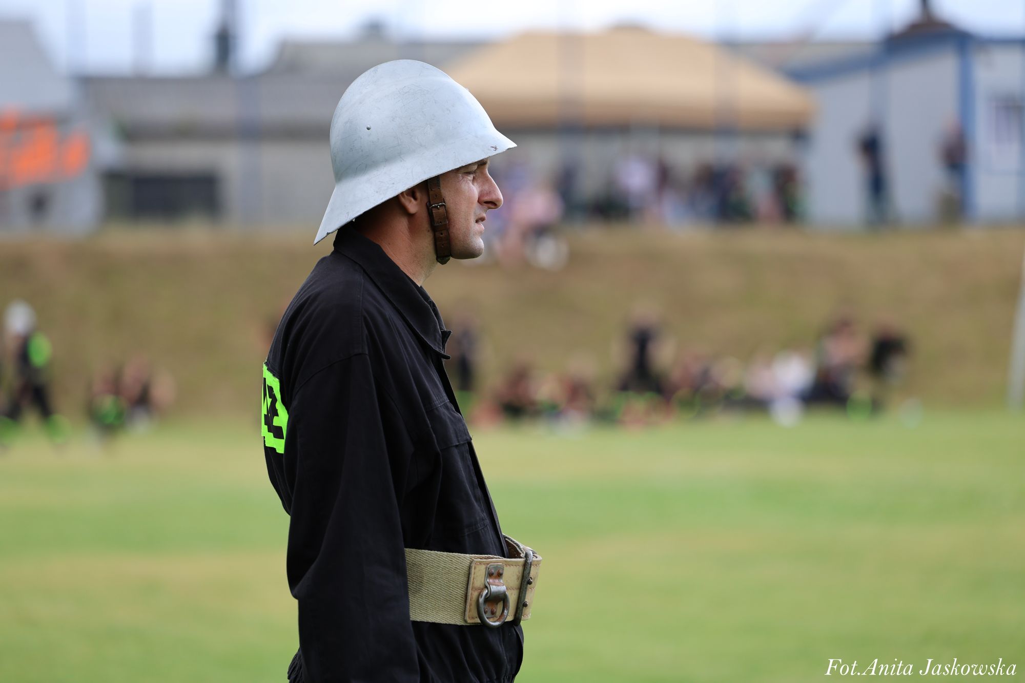 Mężczyzna w czarnym mundurze i białym hełmie na tle zielonej trawy i rozmazanych postaci oraz budynków.