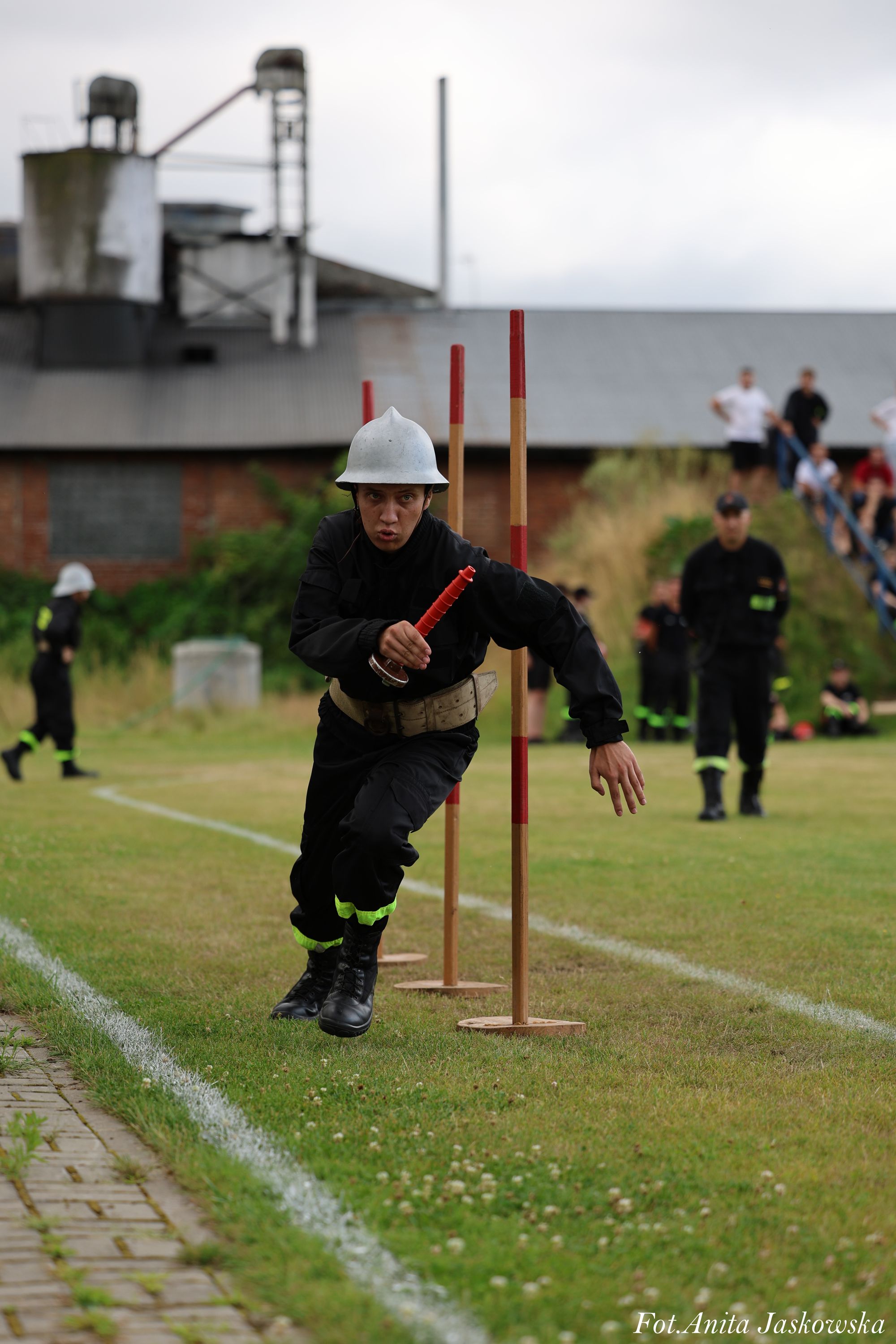 Mężczyzna w czarnym mundurze i srebrnym hełmie biegnie po trawie między trzema drewnianymi słupkami z czerwonymi końcówkami, trzymając czerwony przedmiot. W tle inni ludzie.