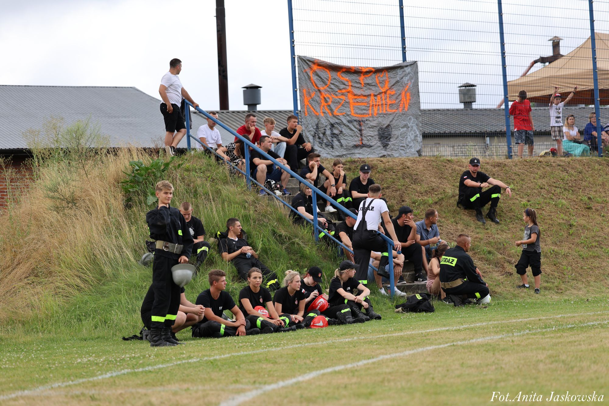 Grupa ludzi w czarnych mundurach z napisem "STRAŻ" siedzi i stoi na trawie oraz schodach, za nimi szary baner z pomarańczowym napisem "OSP KRZEMIEŃ".