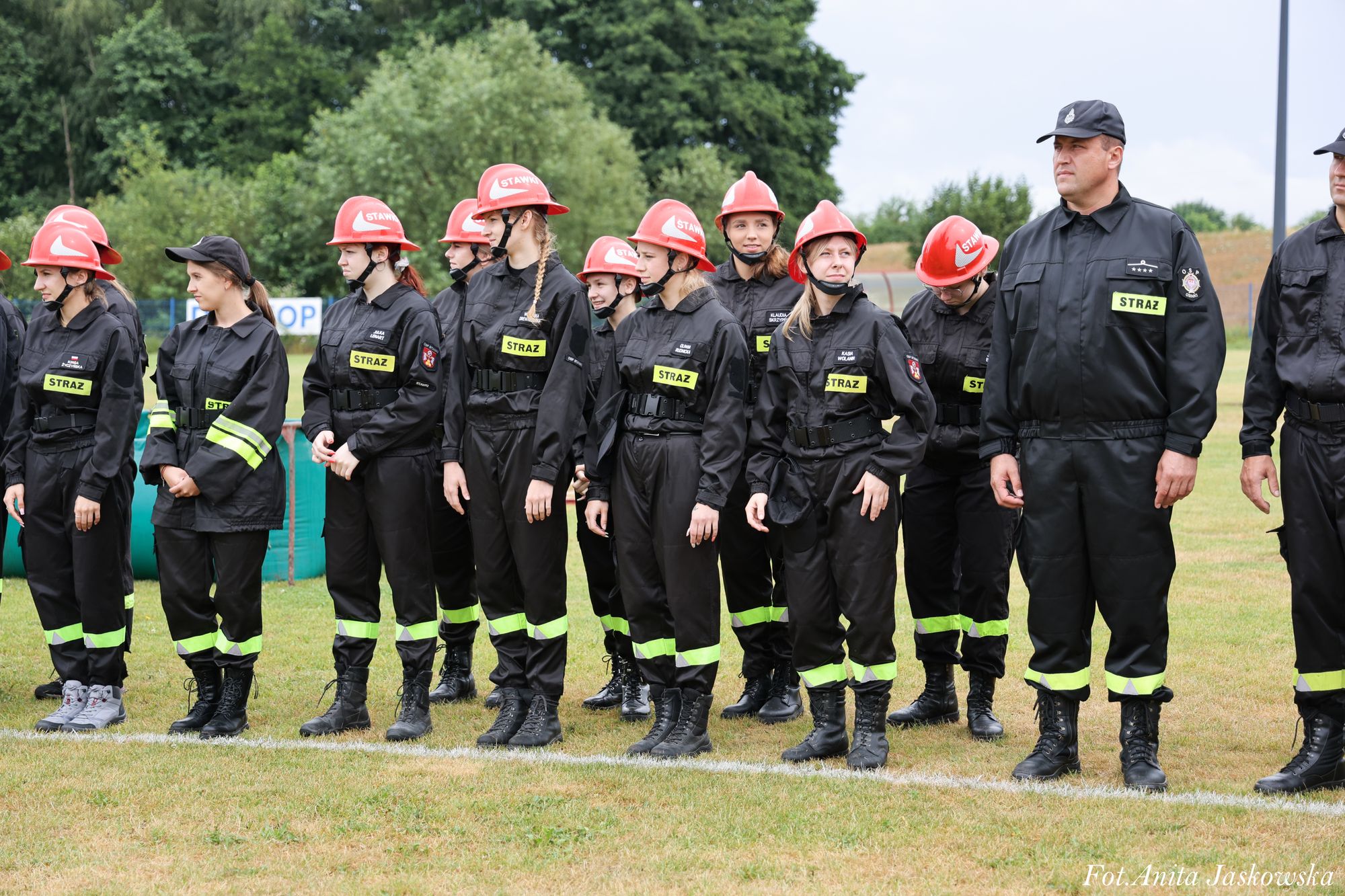 Grupa osób ubranych w czarne mundury z odblaskowymi paskami, czerwone kaski i czarne buty stoi na trawie, w tle drzewa i płot.