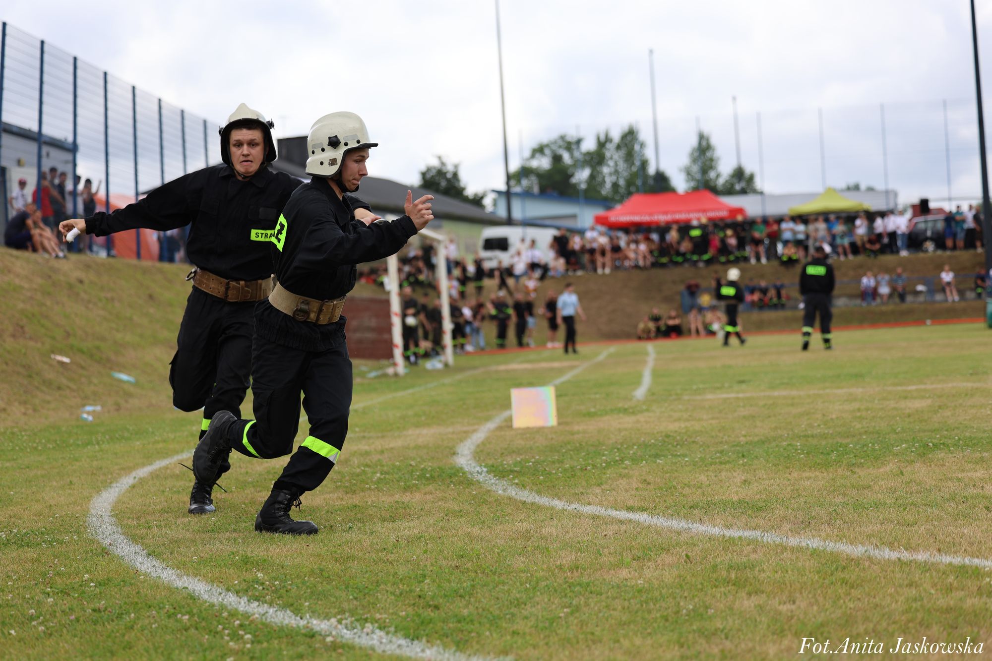 Dwóch strażaków w czarnych mundurach i hełmach biegnie po zielonym boisku z białymi liniami, w tle tłum ludzi na trybunach i kolorowe namioty.