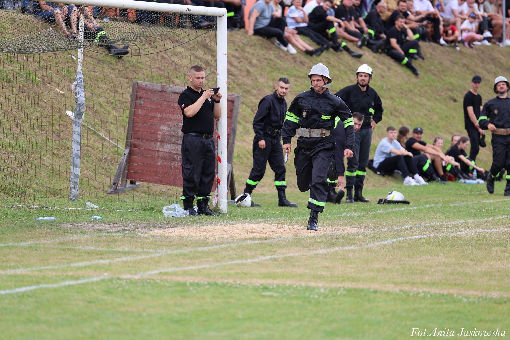 Mężczyzna w czarnym mundurze strażackim z jasnym hełmem biegnie po trawiastej murawie, w tle inni strażacy i widzowie siedzący na skarpie.
