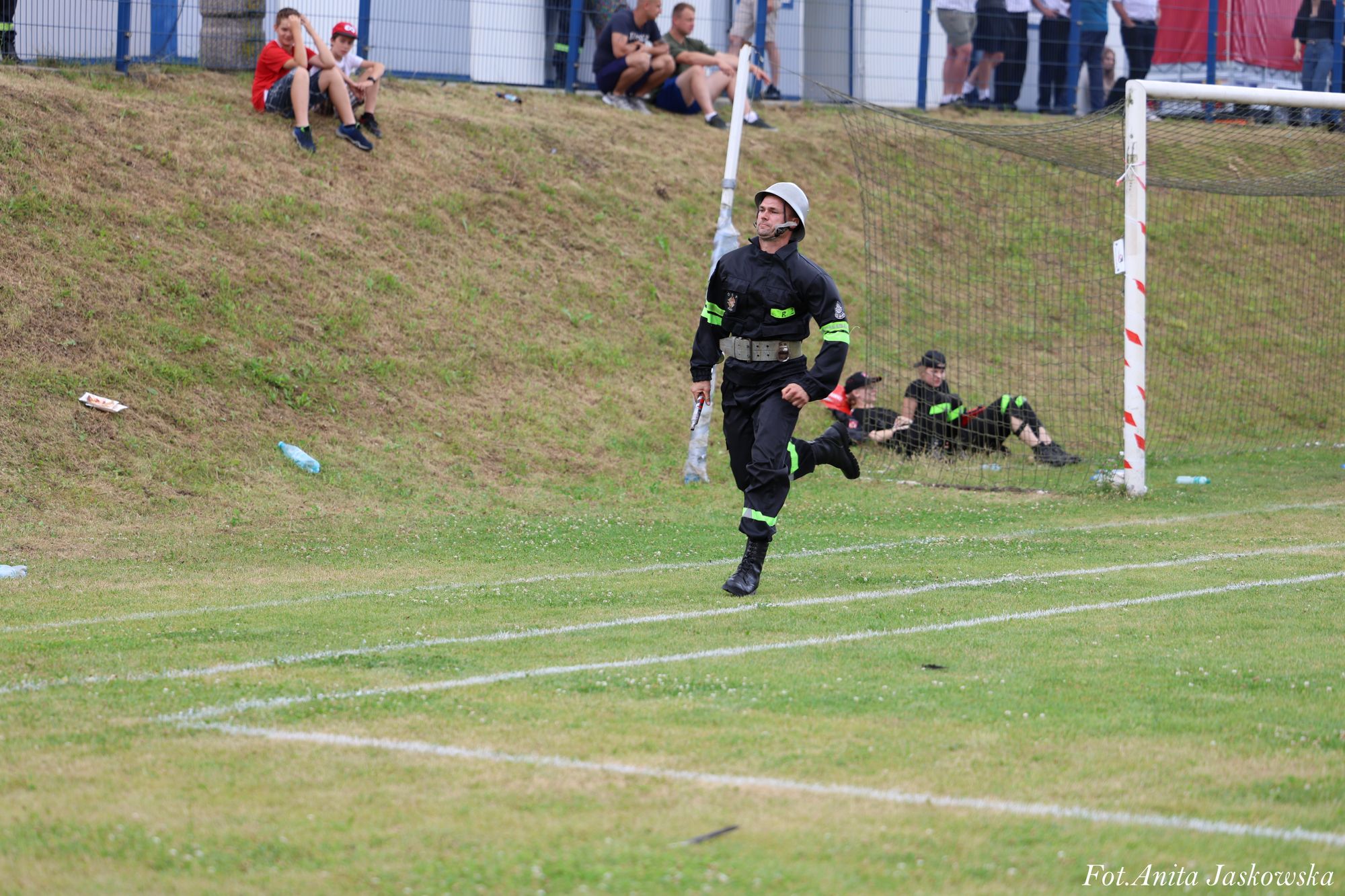 Mężczyzna w czarnym ubraniu roboczym z odblaskowymi zielonymi paskami i białym hełmem biegnie po zielonym boisku z białymi liniami, w tle siedzą i stoją ludzie na skarpie oraz przy bramce.