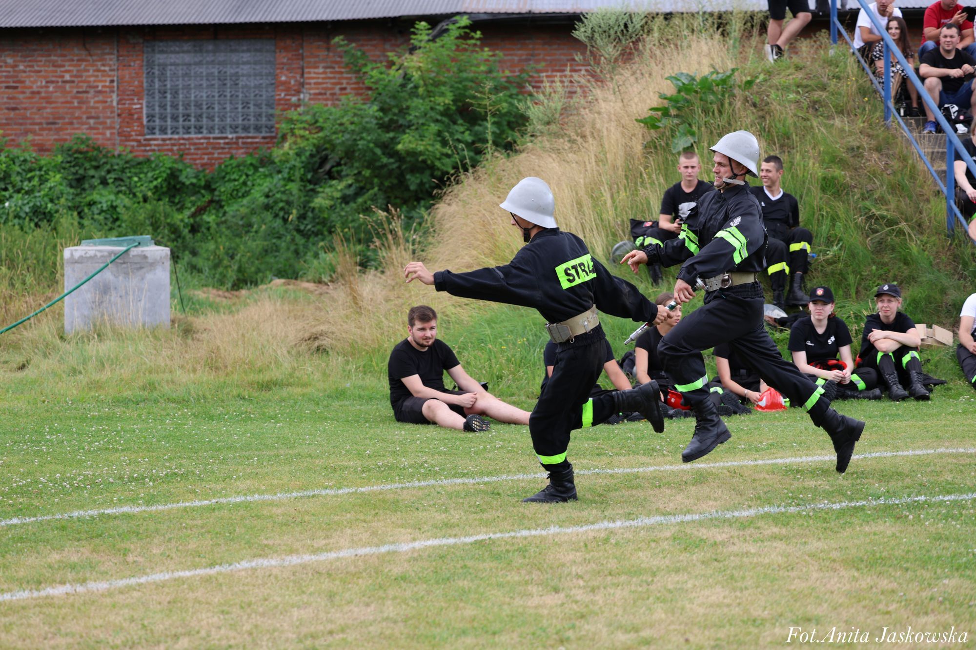 Dwóch mężczyzn w czarnych mundurach ze srebrnymi hełmami i odblaskowymi pasami biegnie po zielonym boisku, w tle grupa osób siedzących na trawie i schodach, ceglany budynek, zieleń.