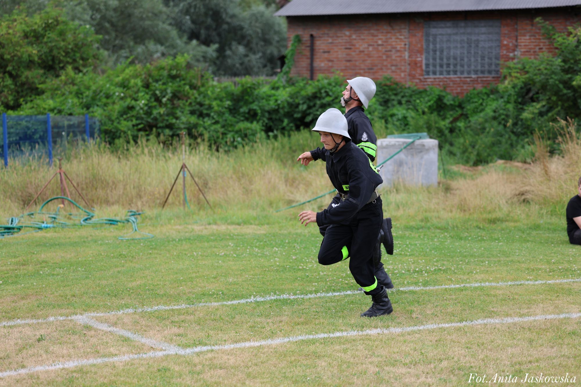 Dwóch mężczyzn w czarnych mundurach i białych hełmach biegnie po zielonym boisku z białymi liniami, w tle zarośla i ceglany budynek.