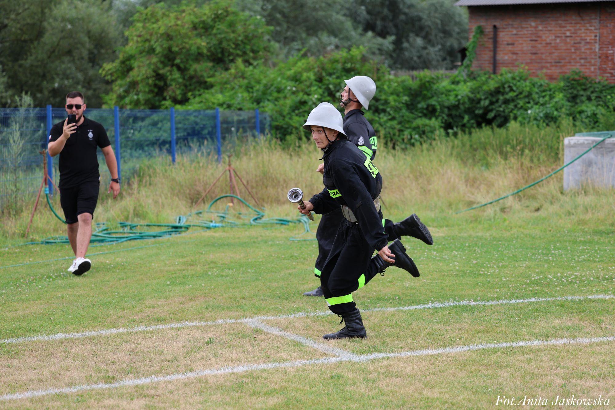 Dwóch mężczyzn w czarnych mundurach z białymi hełmami biegnie po trawiastym boisku, jeden trzyma metalowy element, w tle mężczyzna w czarnym stroju robi zdjęcie telefonem.
