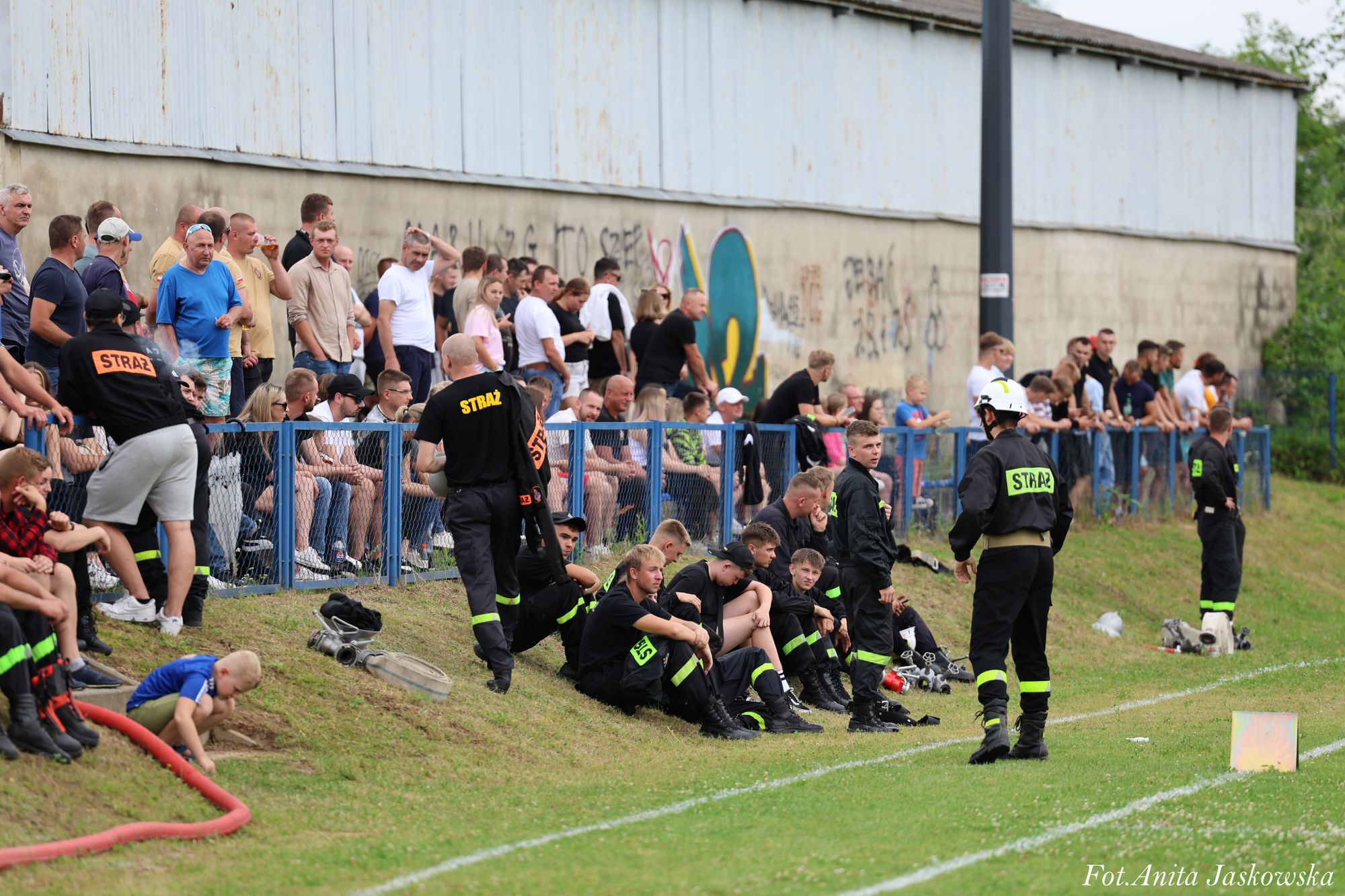 Grupa strażaków w czarnych mundurach z żółtymi napisami "STRAŻ" siedzi i stoi na trawie przy boisku sportowym, za nimi stoi ogrodzenie i tłum ludzi na tle ściany z graffiti.