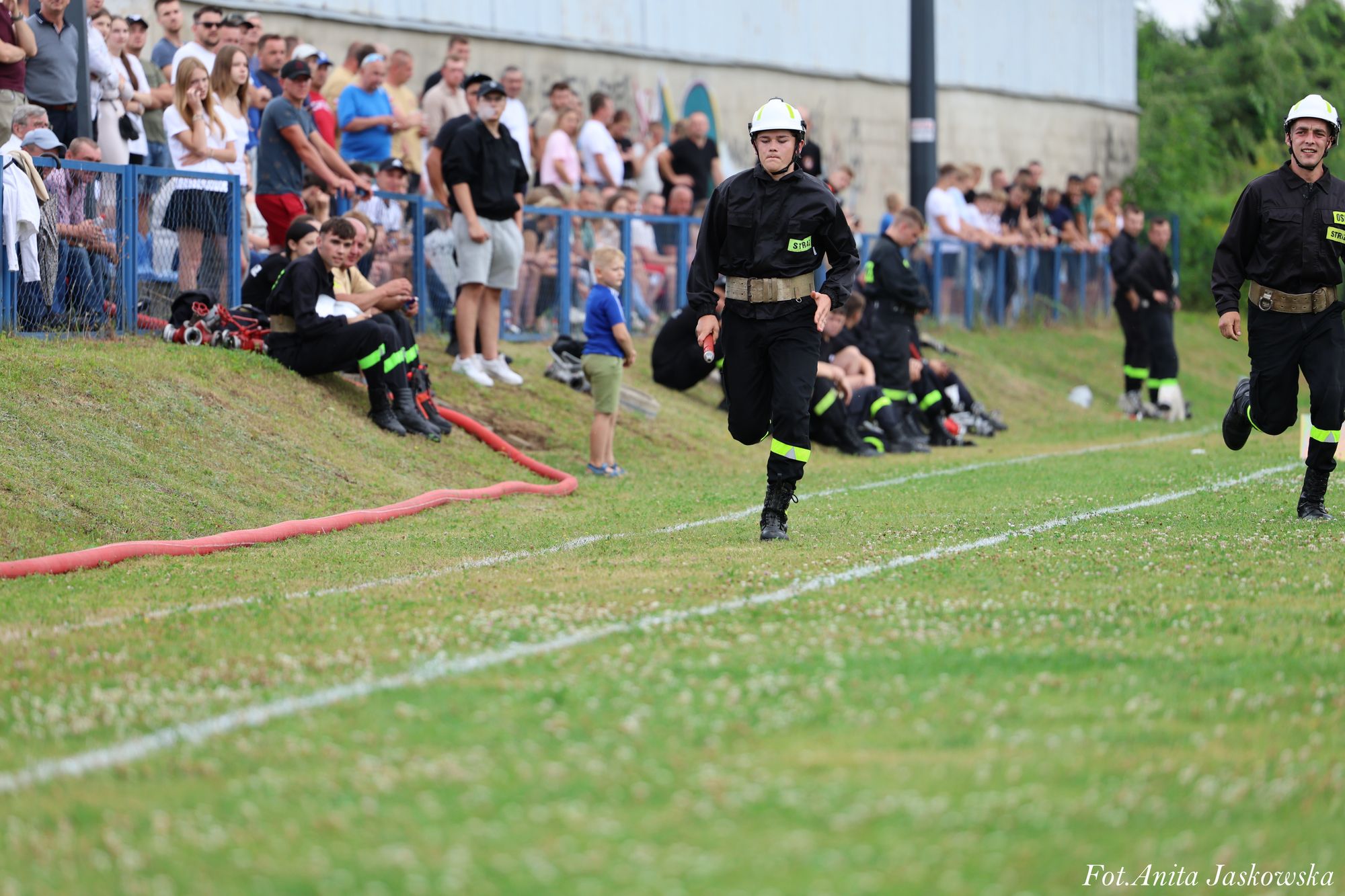 Dwóch strażaków w czarnych mundurach i białych hełmach biegnie po zielonym boisku, w tle tłum ludzi za niebieskim ogrodzeniem.