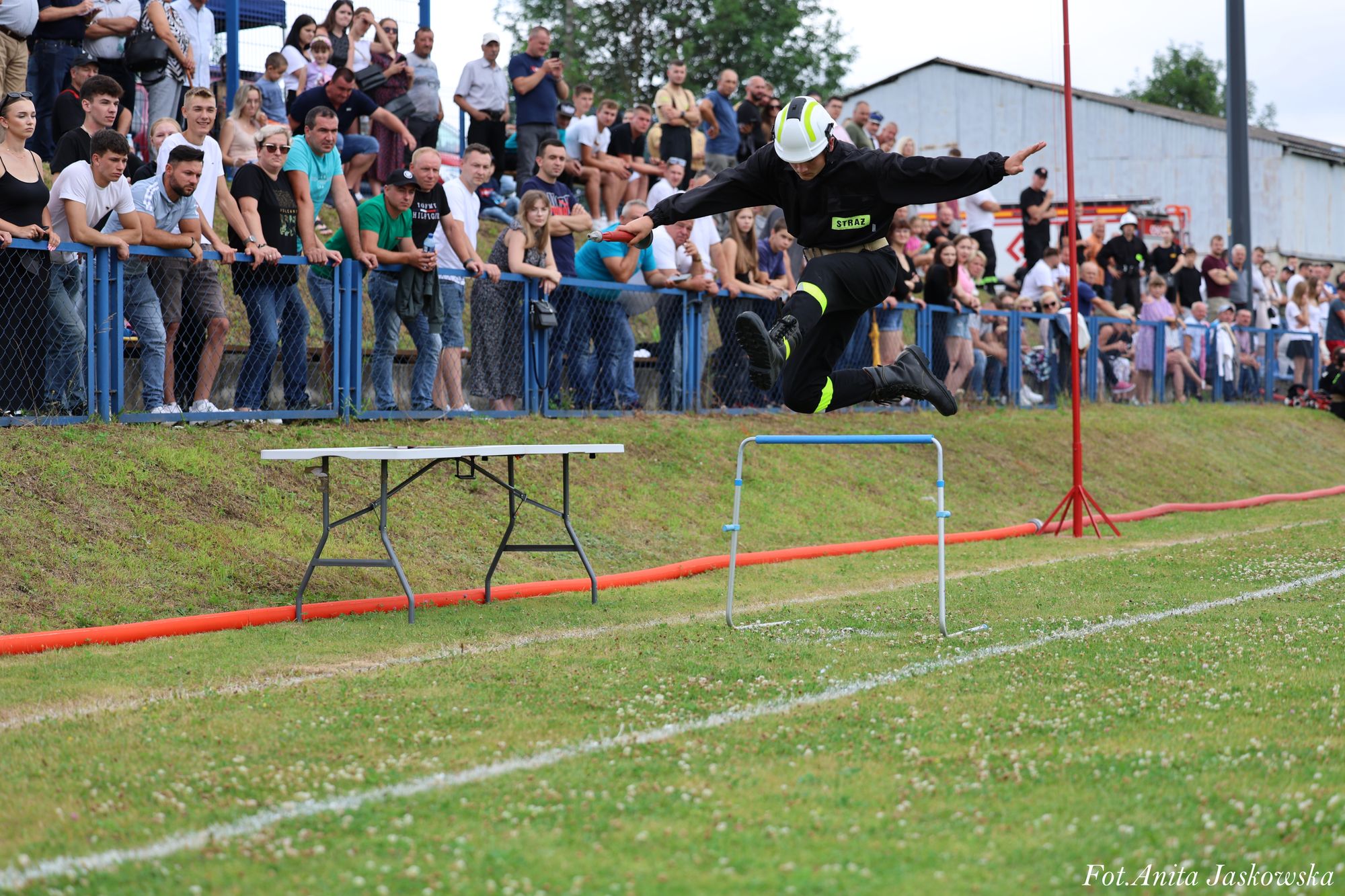 Strażak w czarnym ubraniu z odblaskowymi elementami i białym hełmem skacze nad niebieską przeszkodą na zielonym boisku, w tle tłum ludzi za niebieskim ogrodzeniem.