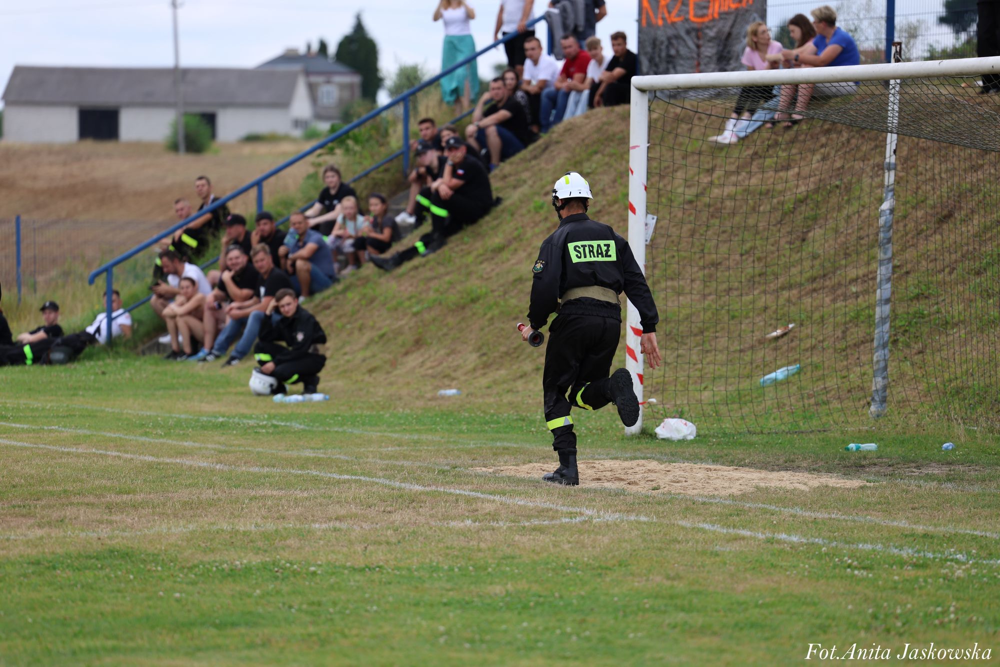 Strażak w czarnym mundurze i białym kasku biegnący obok bramki na trawiastym boisku, w tle siedzący ludzie na zielonym wzniesieniu i trybunach.