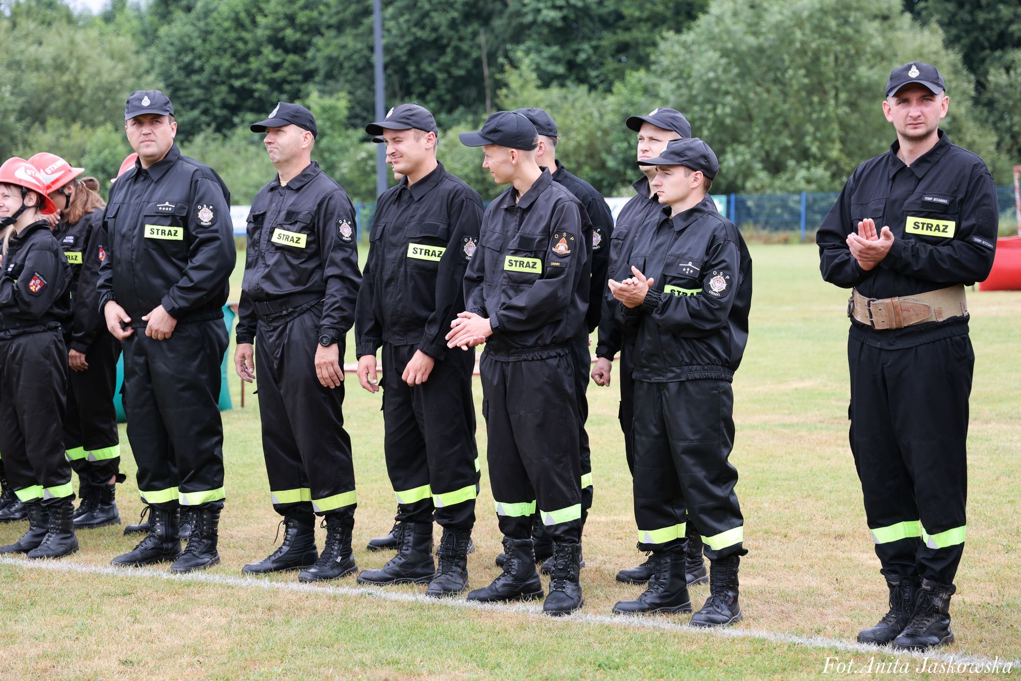 Grupa osób w czarnych mundurach strażackich z żółtymi napisami "STRAŻ" stoi na trawie, w tle drzewa i ogrodzenie. Niektórzy klaszczą.