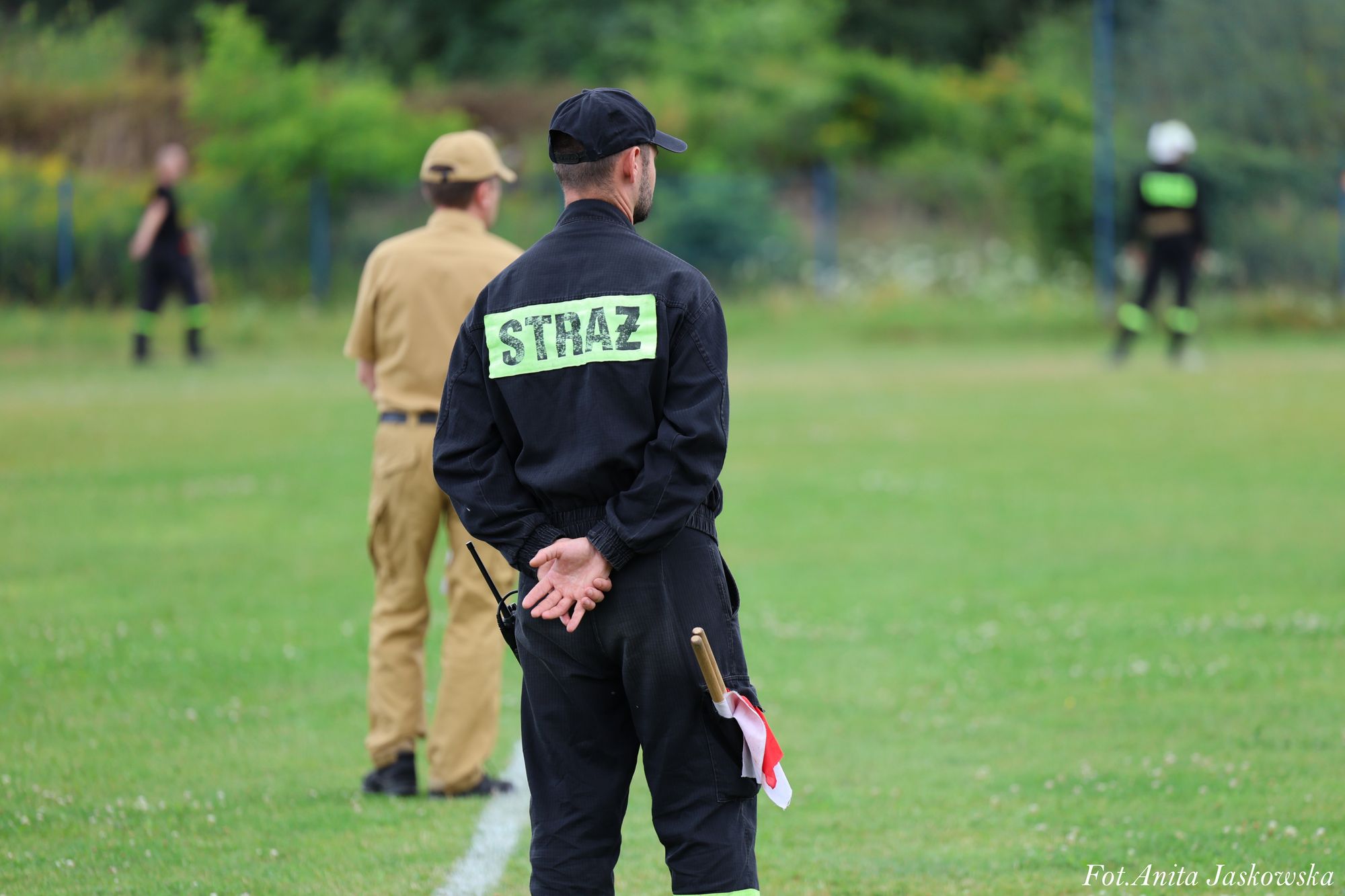 Mężczyzna w czarnym mundurze z napisem "STRAŻ" na plecach, trzymający za sobą chorągiewkę, na zielonym boisku, w tle dwie inne postaci w mundurach.