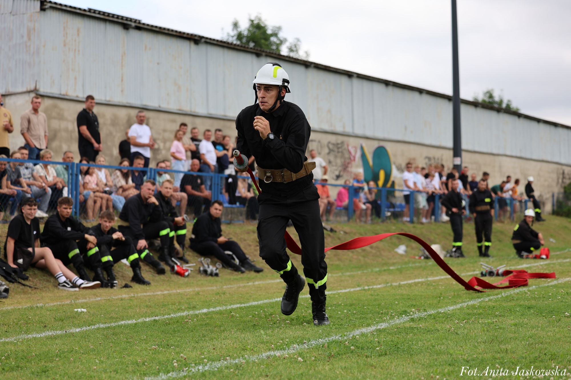 Mężczyzna w czarnym uniformie i białym hełmie biegnie z czerwoną wężem strażackim na zielonym boisku, w tle siedzą i stoją ludzie przy ogrodzeniu.