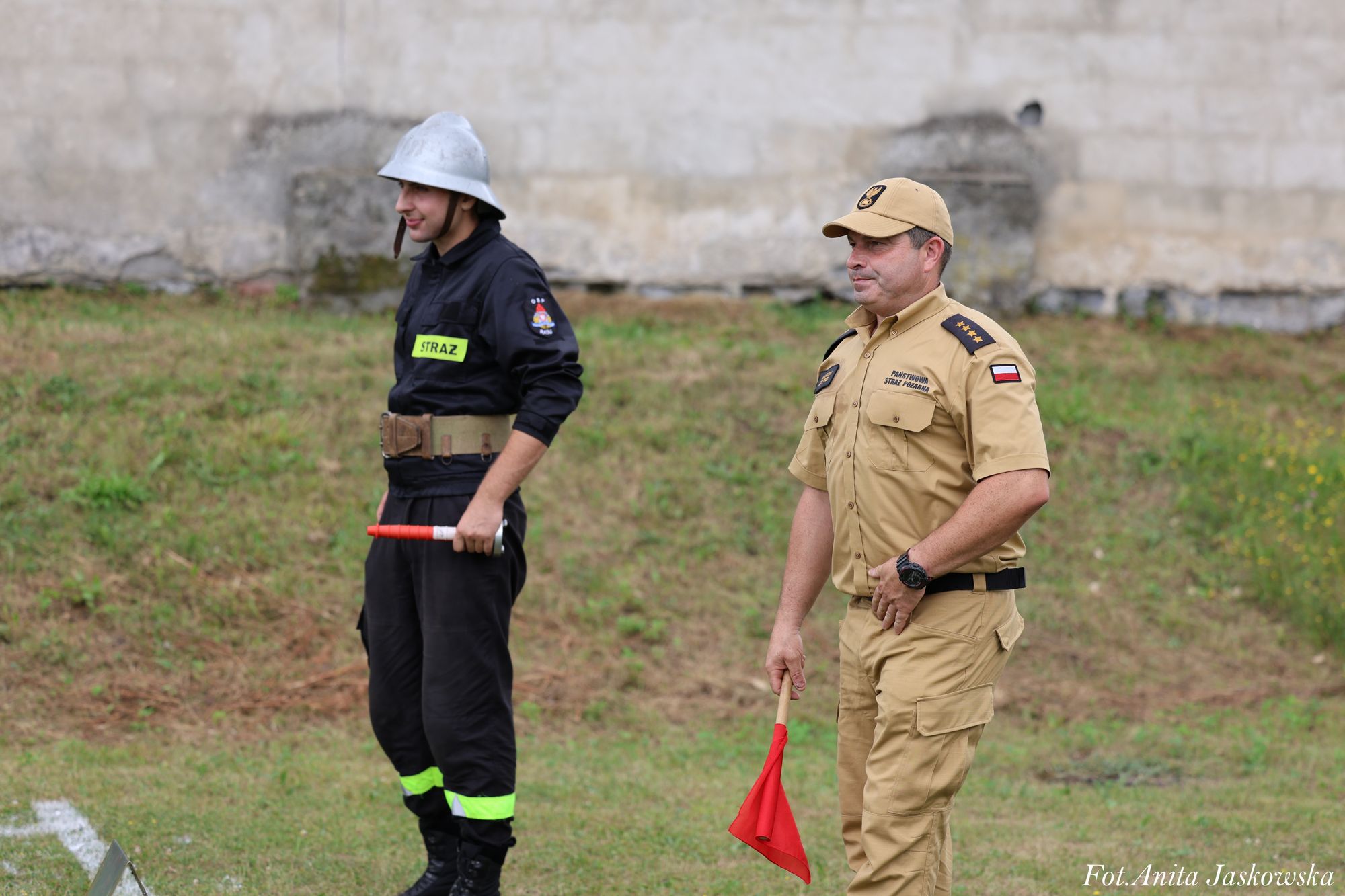 Dwaj mężczyźni w mundurach strażackich stoją na trawie, jeden w czarnym kombinezonie z napisem "STRAŻ" i hełmem, drugi w beżowym mundurze z czapką, trzyma czerwony trójkątny znak.